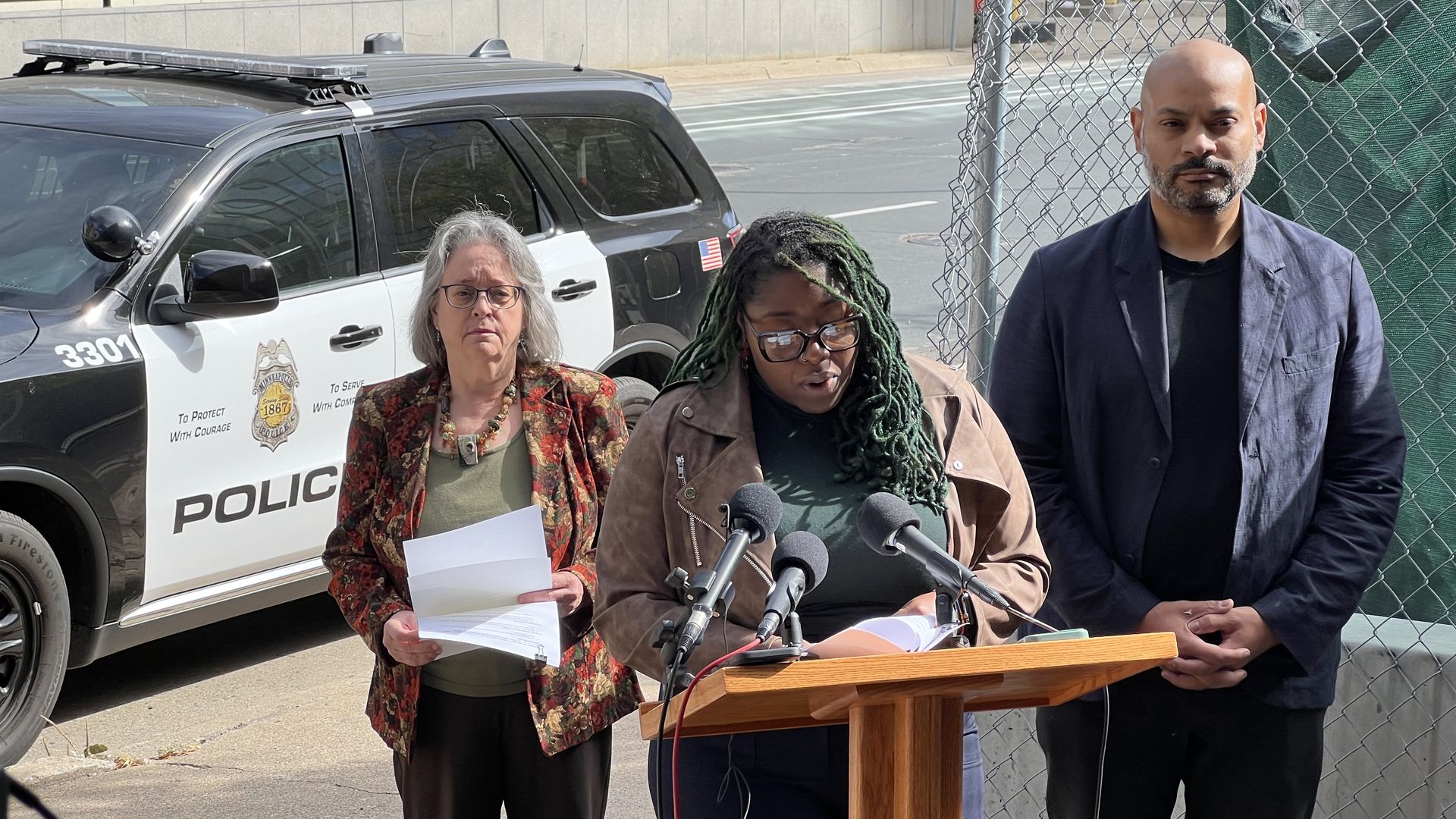 Three people stand at a podium for a press conference in front of a black-and-white police SUV