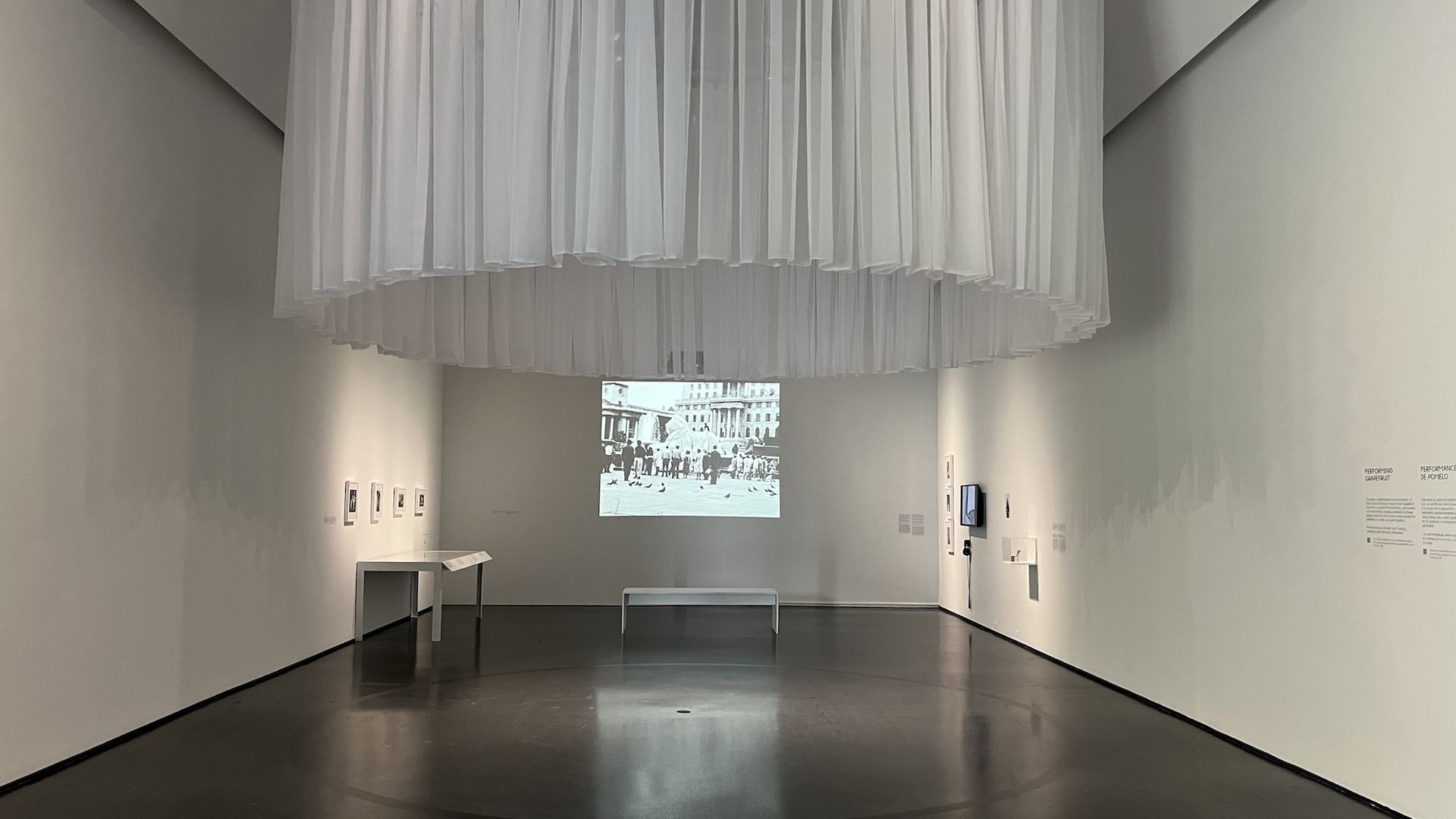 Minimalist gallery room with white walls and floor. A white bench faces a black-and-white photo projected on the far wall. White sheer curtains hang from the ceiling forming a circular shape.