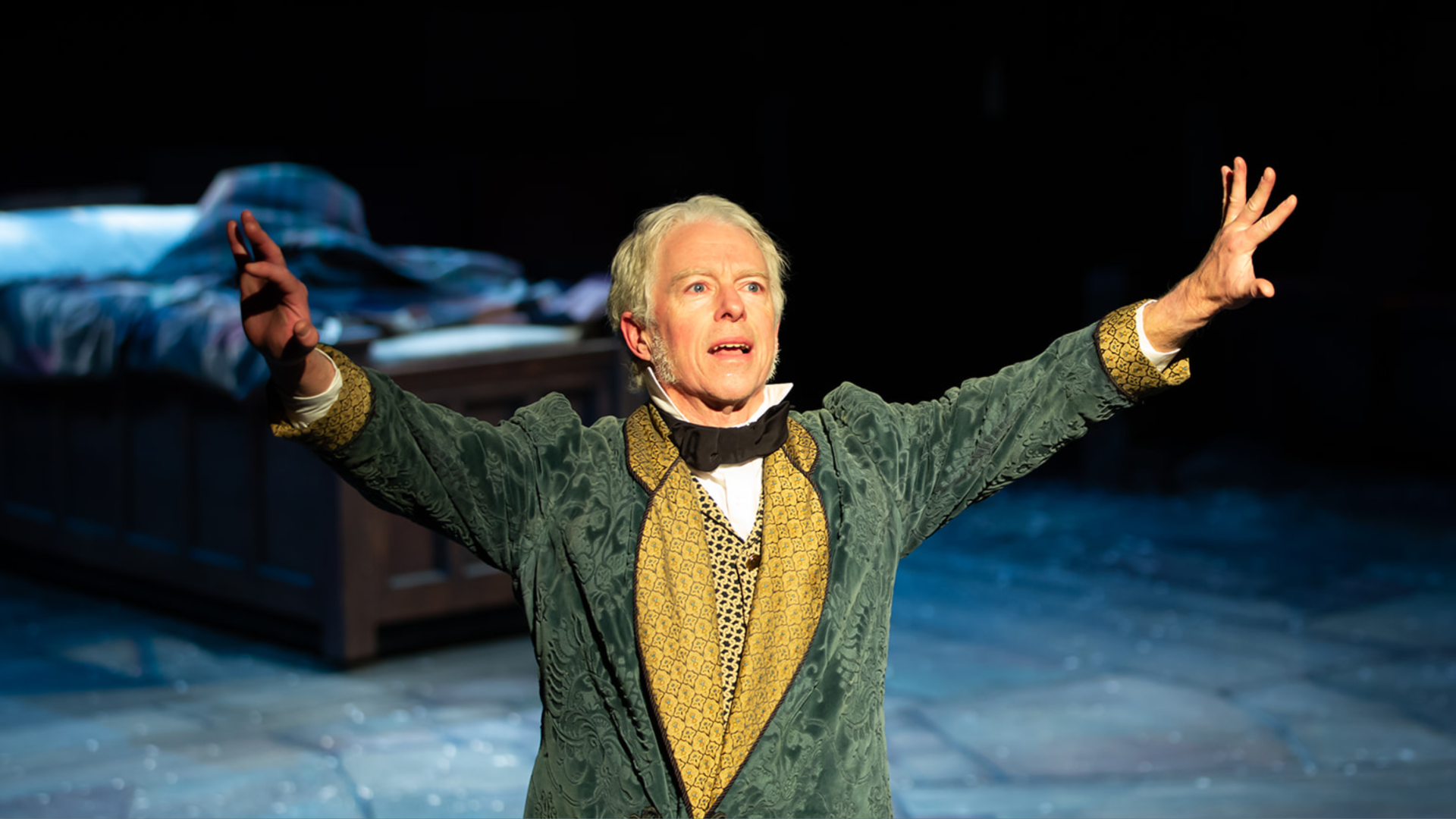 A man dressed in green 19th-century looking garb holds his hands up in the air, looking out, with a bed in the background behind him. 