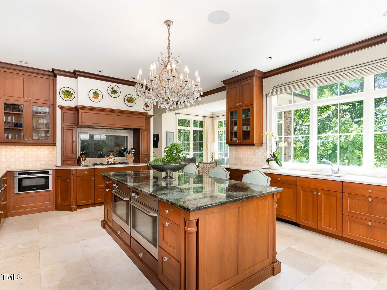 Bright kitchen with wood cabinets, large island with dark stone countertop, chandelier, wall plates as decoration, and large window showing green trees outside.