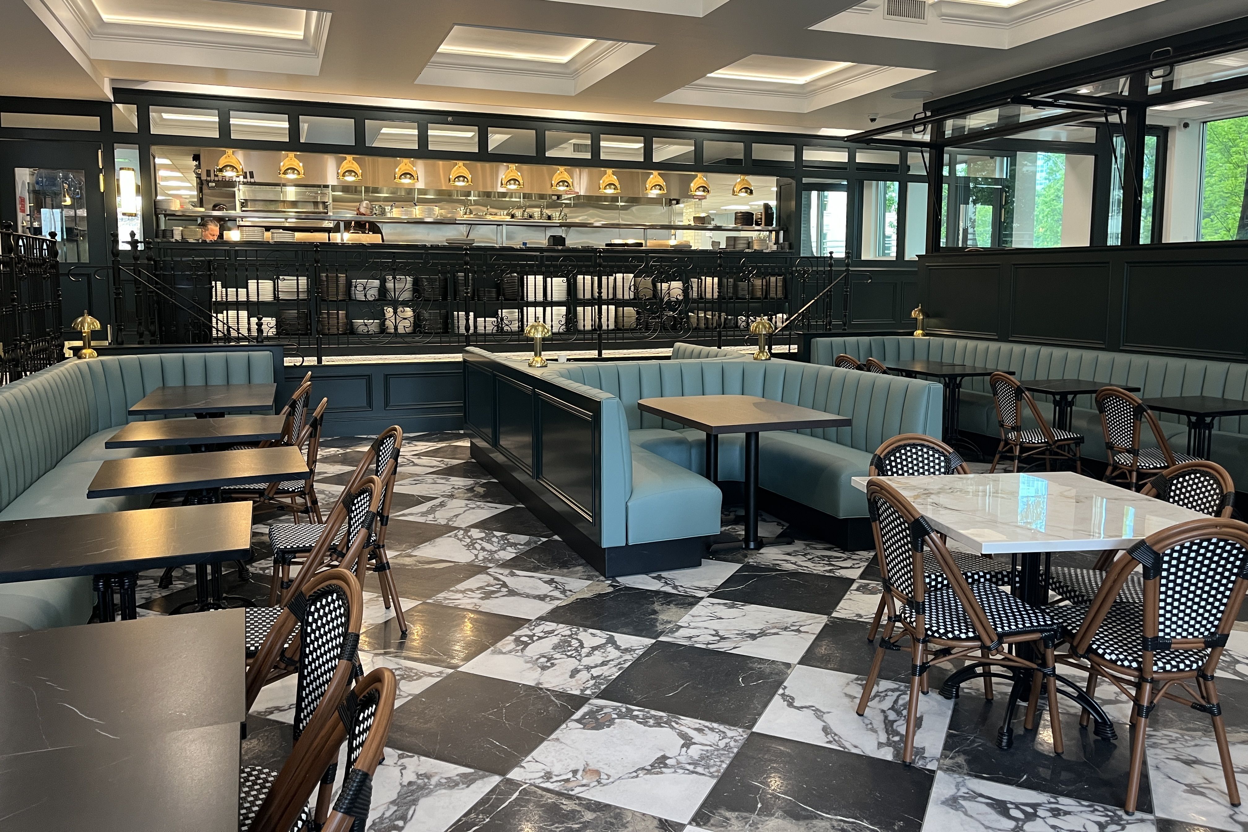 Inside a stylish restaurant with an open kitchen behind glass, gold pendant lights, dark teal paneling, blue upholstered booths, and marble checkered floors with wicker chairs.