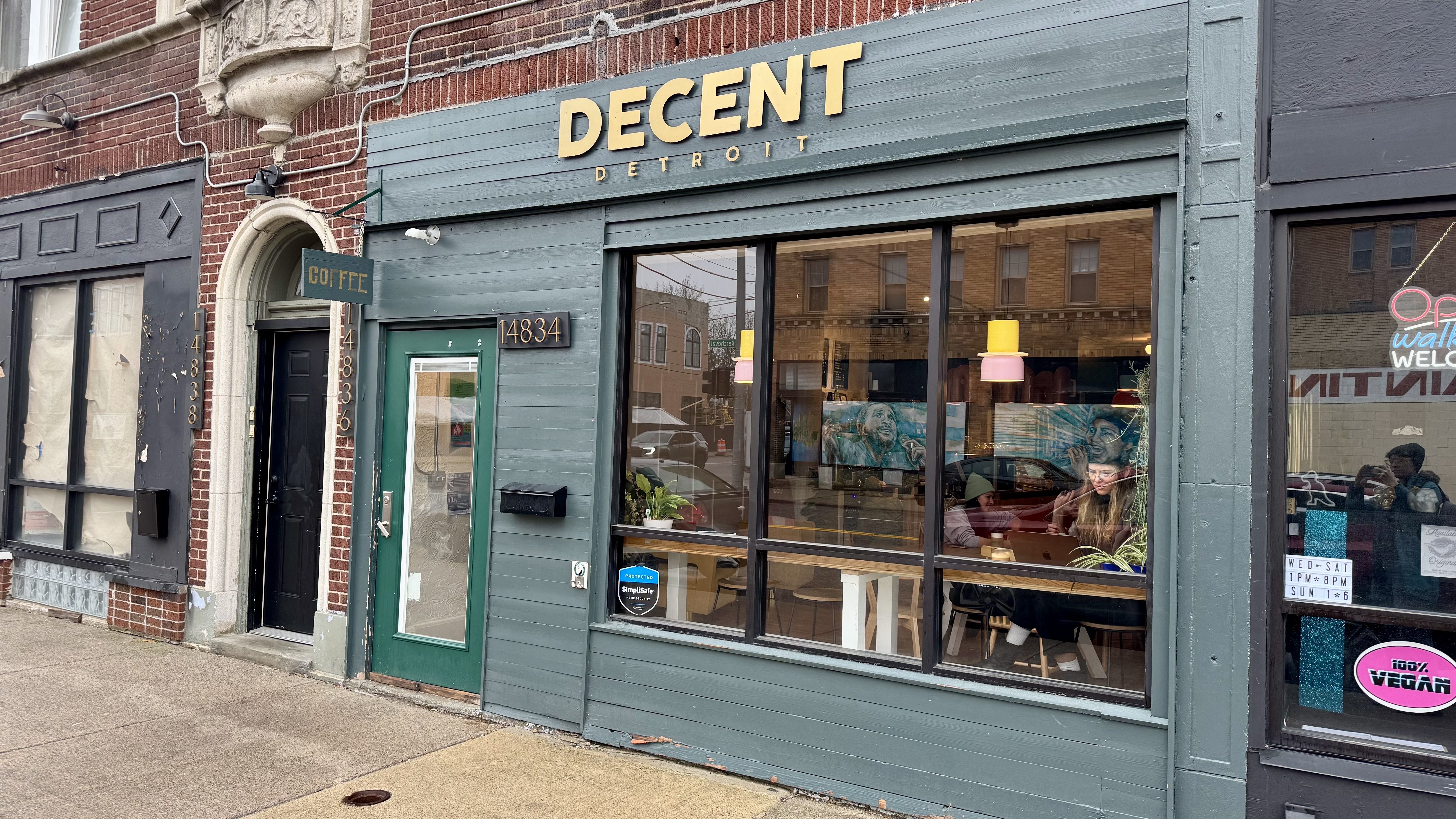 Exterior of Decent Detroit coffee shop with gray-green facade and large window showing people inside.