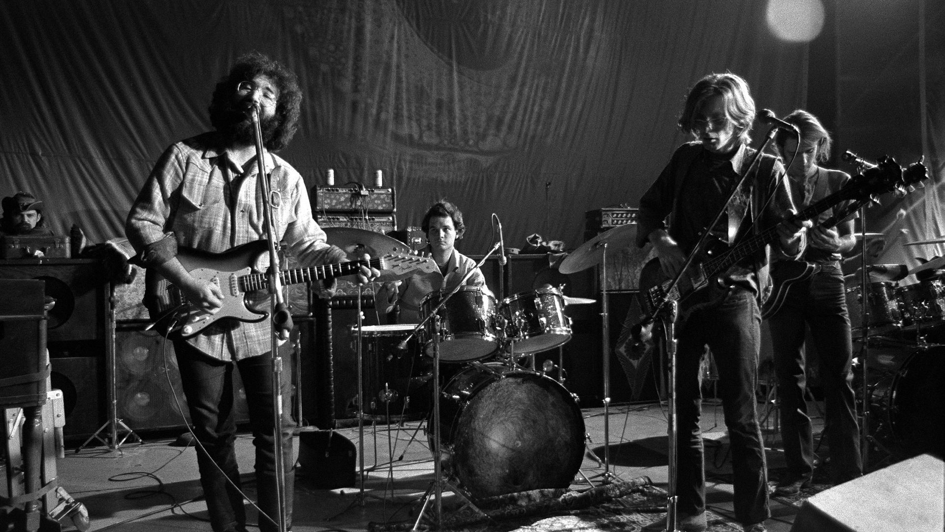 Black-and-white photo of a live rock band performing on stage: left, a bearded guitarist-singer sings into a mic; center drummer; right, another guitarist/vocalist at a second mic, with amps and rug visible.