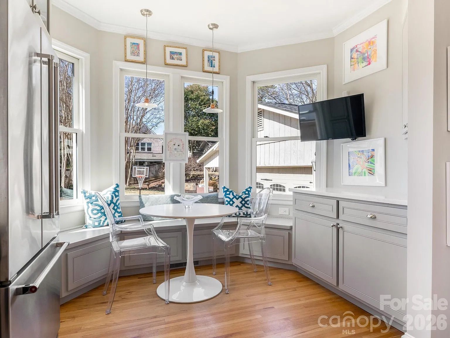 Bright kitchen nook with large windows showing outdoor view, white round table, clear chairs, built-in gray bench with blue patterned pillows, light hardwood floor, and wall-mounted TV.