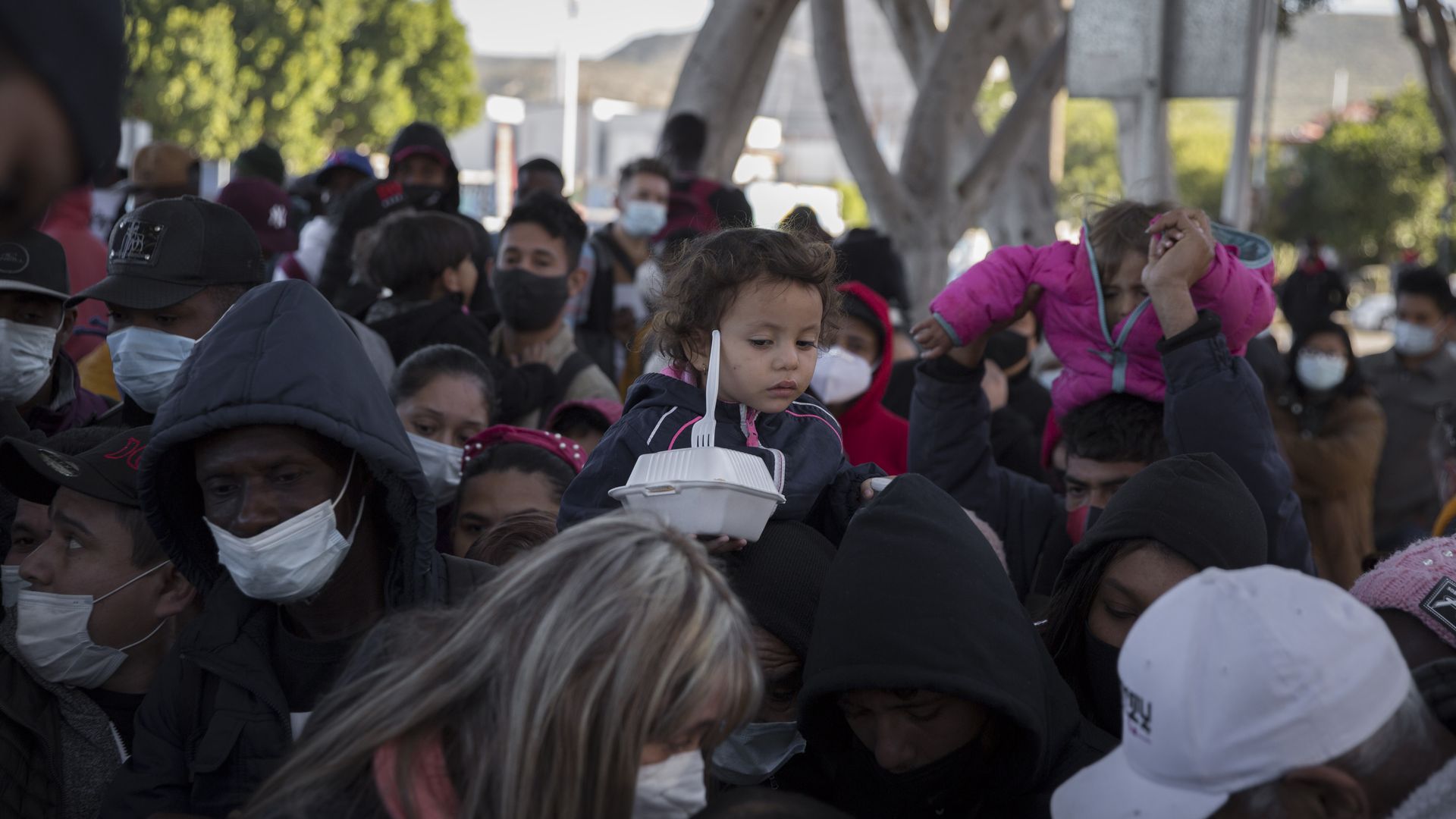 Children are carried on shoulders as scores of asylum seekers wait for a meal and a drink at the "El Chaparral" border crossing