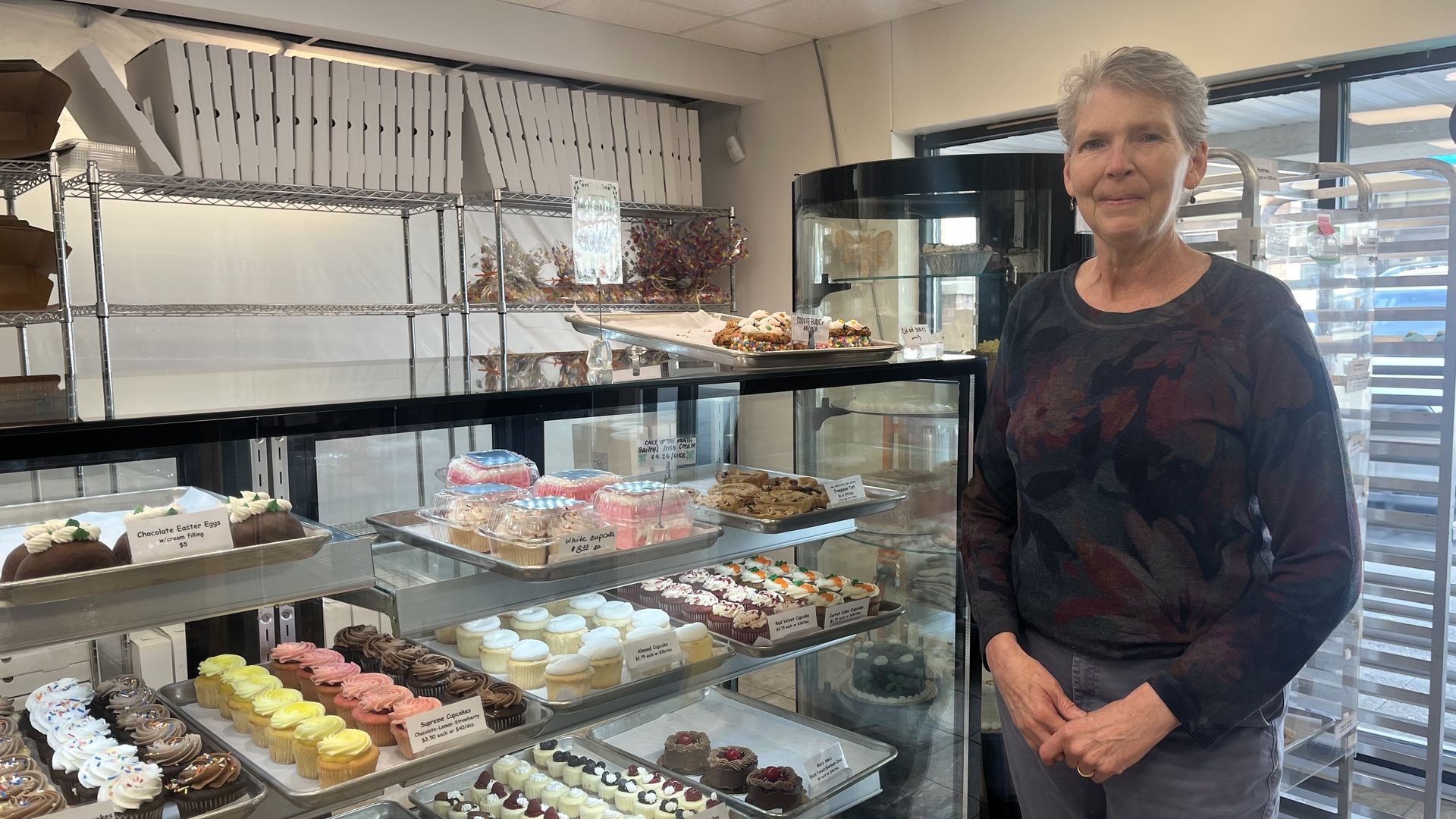 Smiling older woman with short gray hair and a dark floral shirt stands beside a glass bakery display filled with cupcakes and pastries, with metal shelves and boxes in the background.