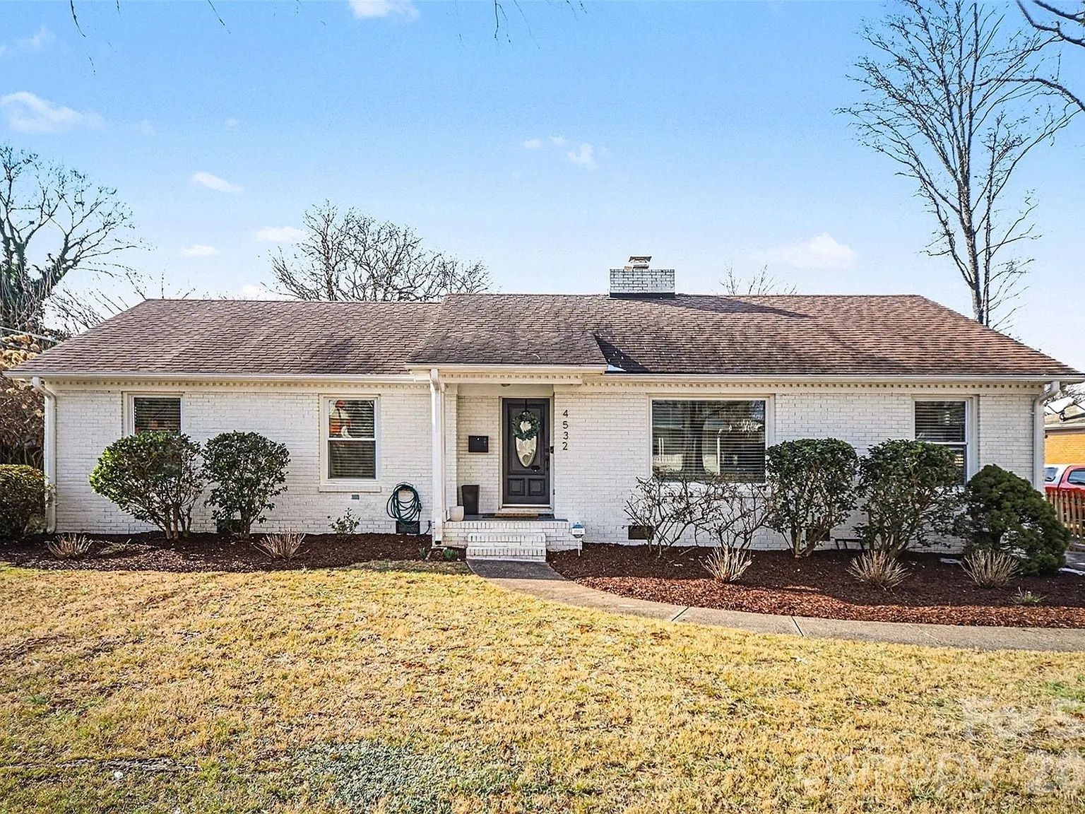 Single-story white brick house with a brown shingled roof, black front door, and trimmed bushes in front yard with yellowish grass under a clear blue sky.