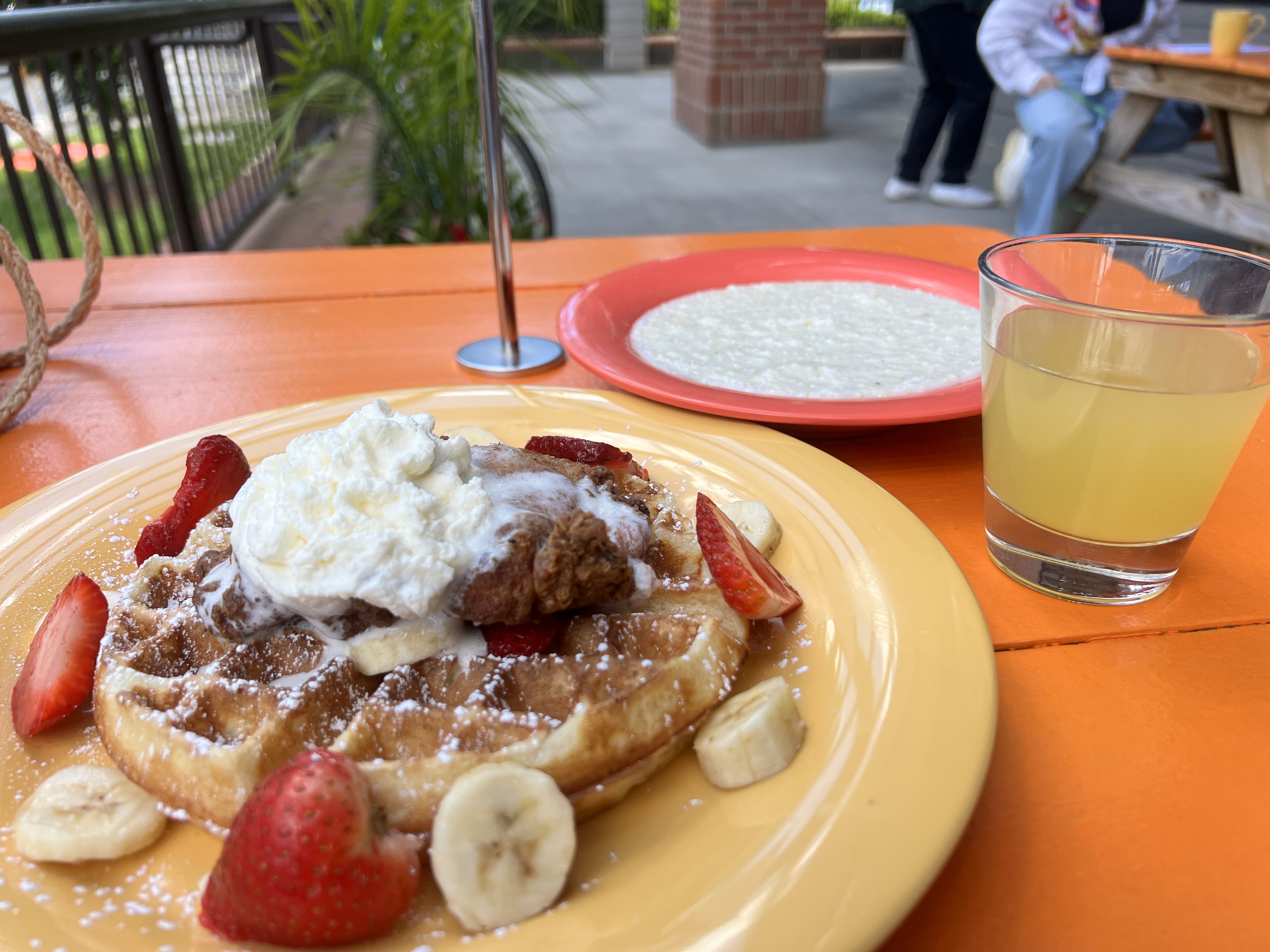 A waffle with whipped cream on a yellow plate, surrounded by banana slices and strawberries. A glass of pale lemonade and a red plate of oatmeal sit on a bright orange table outdoors.