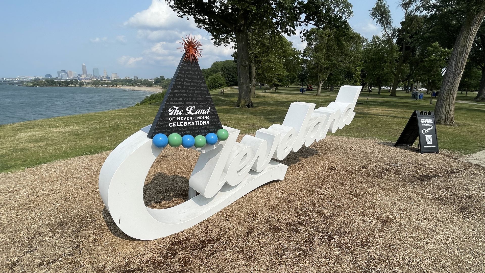 Lakeside park with a large white cursive sculpture spelling "Cleveland" beside a black pyramid inscribed with "The Land of Never-Ending Celebrations" topped by a red spiky star; blue and green balls line the base, with trees, water, and distant skyline.
