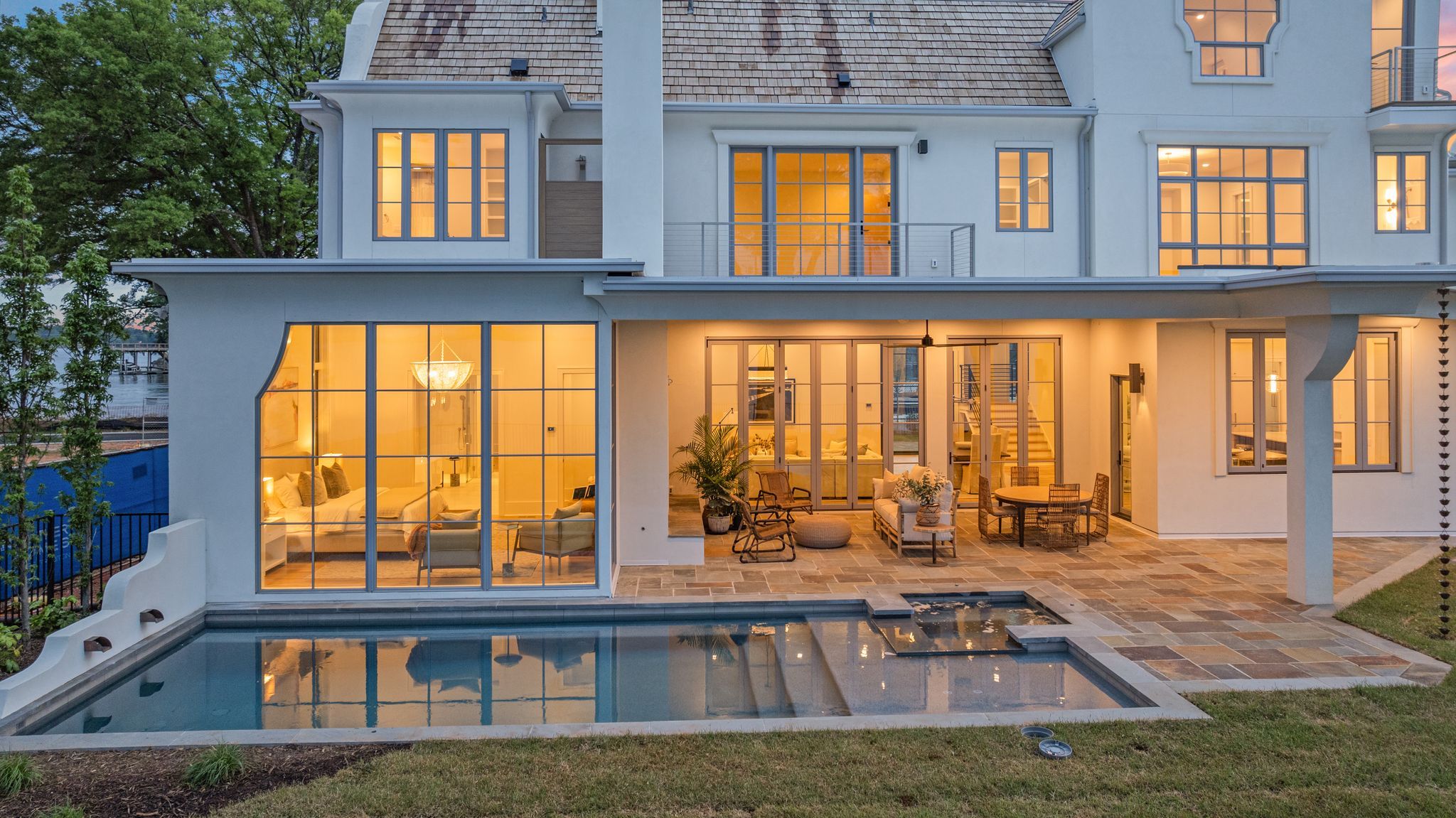 Modern two-story white house with large glass windows glowing warm light. Outdoor patio with seating and a pool reflecting the house and trees at dusk.