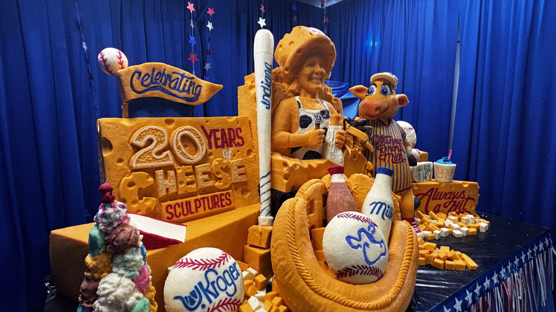 Colorful cheese sculpture display on a blue backdrop, featuring an orange-clad figure among oversized cheese blocks, a striped cow mascot, baseballs, and the sign "20 YEARS OF CHEESE SCULPTURES".