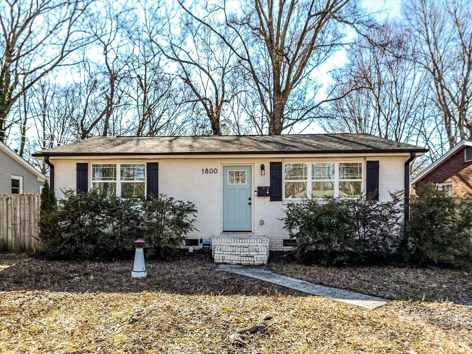 Single-story white brick house with black shutters, light blue front door numbered 1800, surrounded by leafless trees and green bushes, under a clear blue sky.