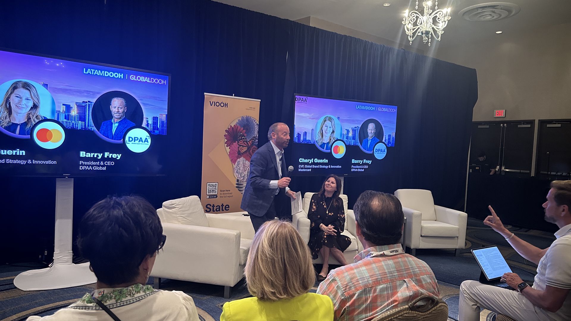Barry Frey standing next to Cheryl Guerin who is seated in a white chair next to him, as four people sit and listen