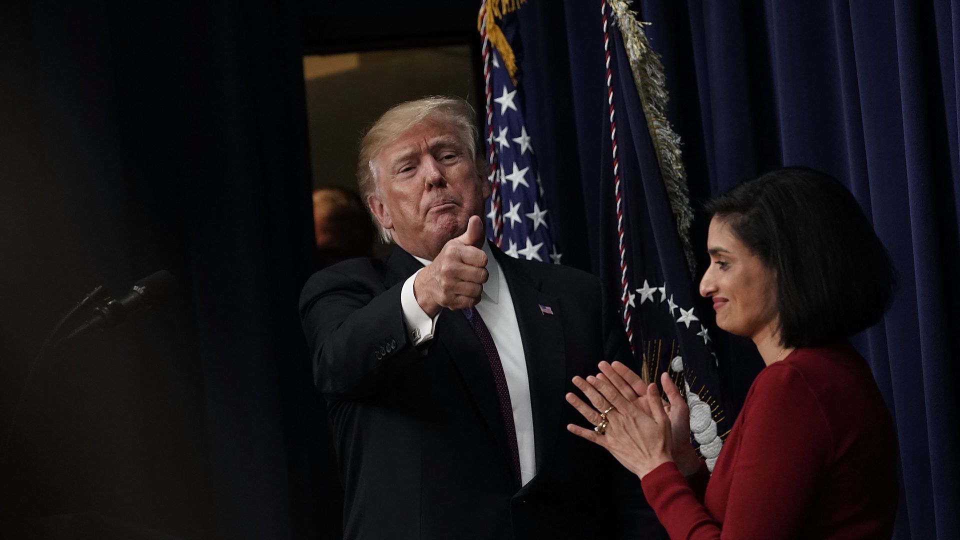 In this image, Trump gives a thumbs up over the CMS administrator, who is clapping. American flag is behind both of them. 