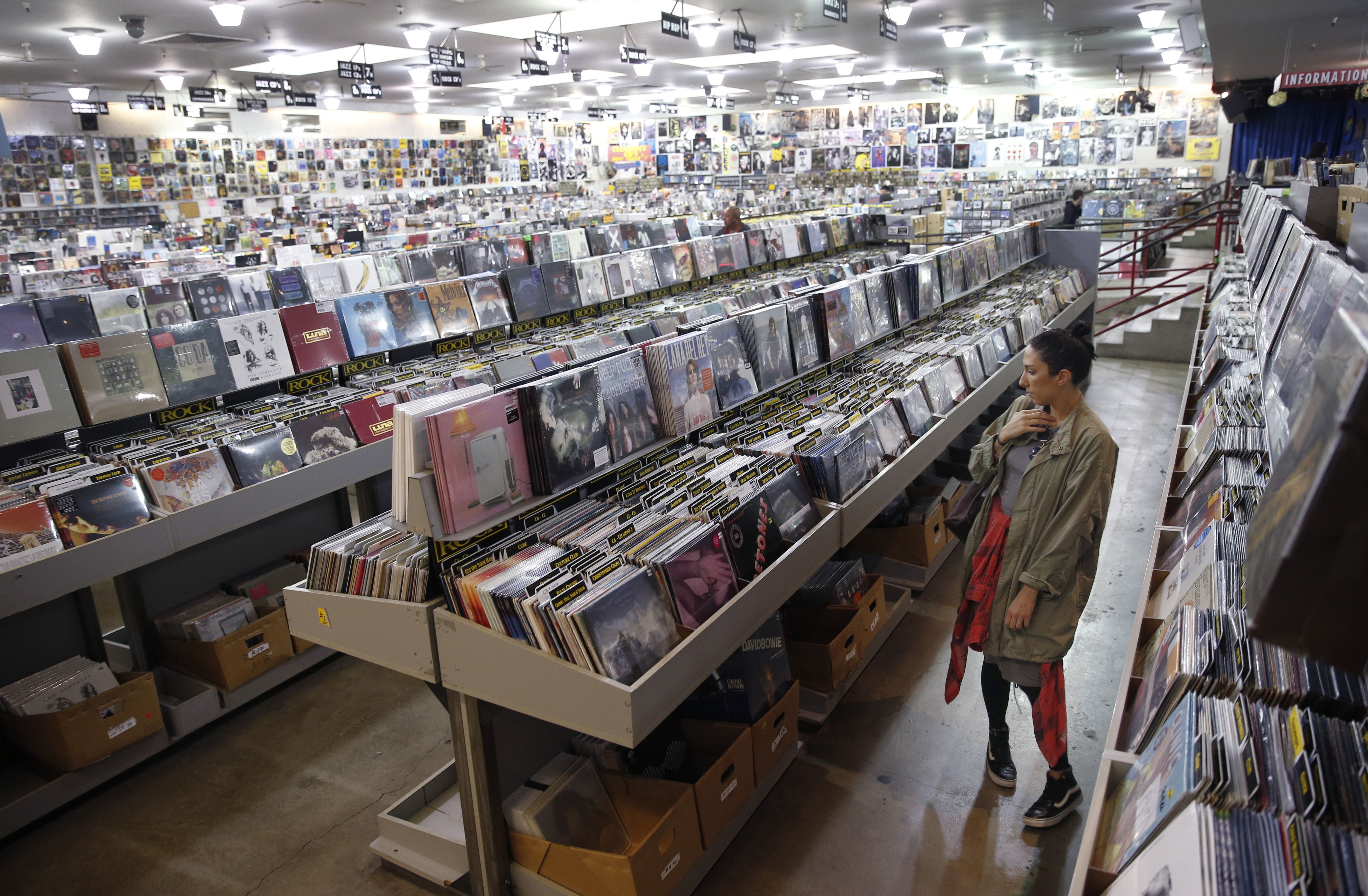 A woman browses vinyl and CD records in a large music store with rows of albums labeled by genre under bright ceiling lights.