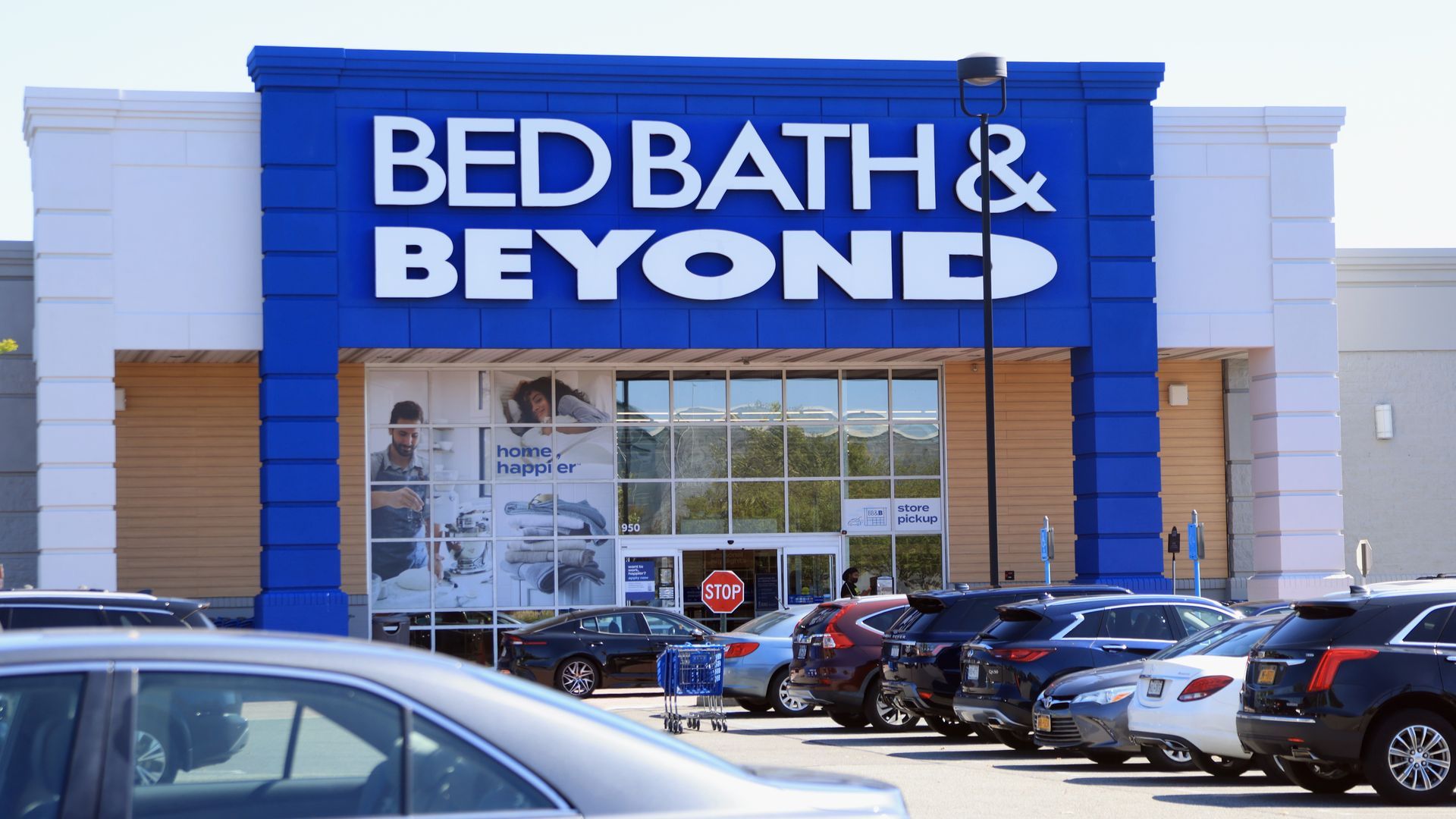 A Bed Bath & Beyond store front, in blue and white, with a parking lot filled with cars.