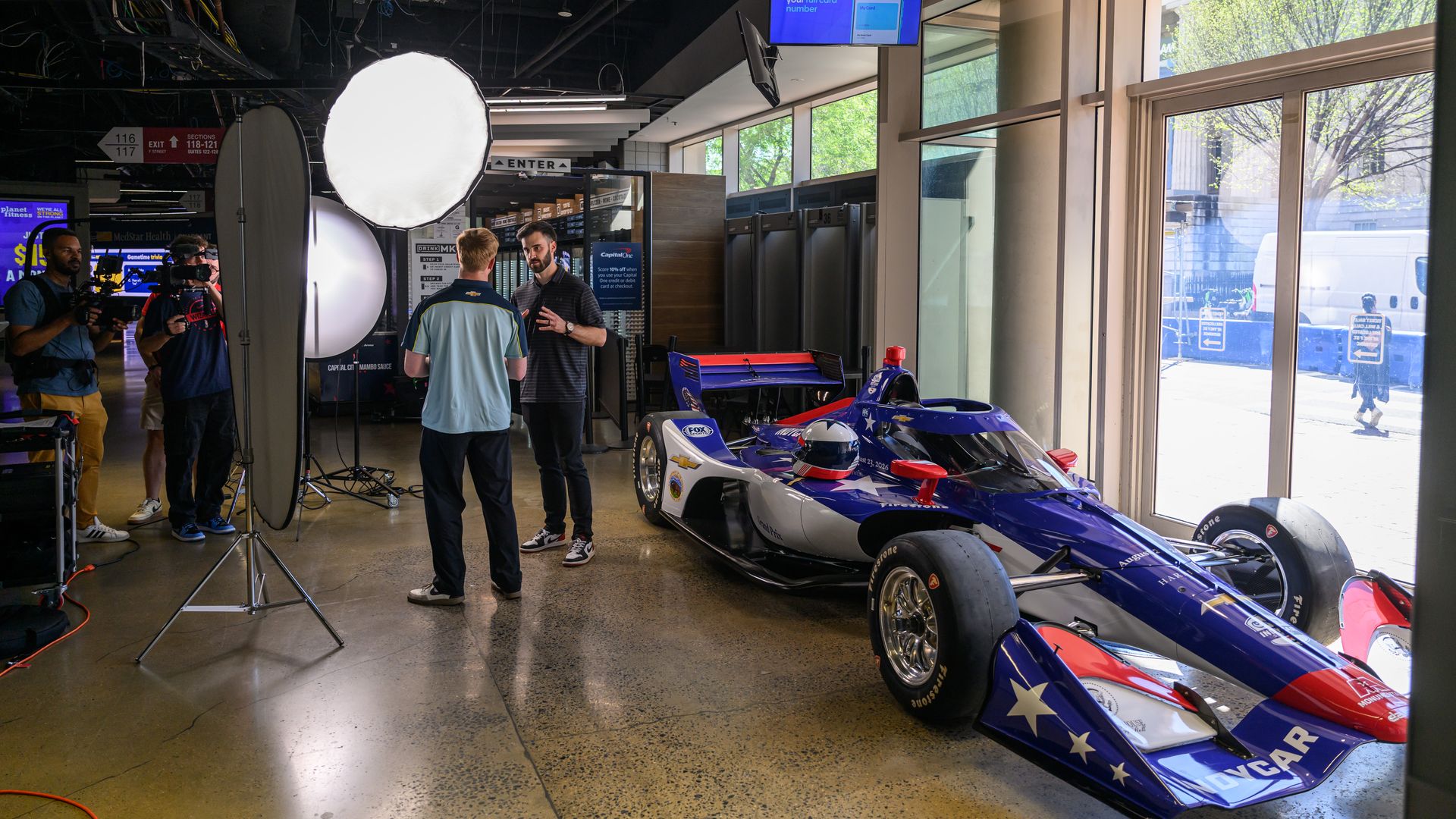 Indoor studio with a blue, white, and red race car on display near big windows; film crew with cameras and a circular softbox light, two men talking beside the car.