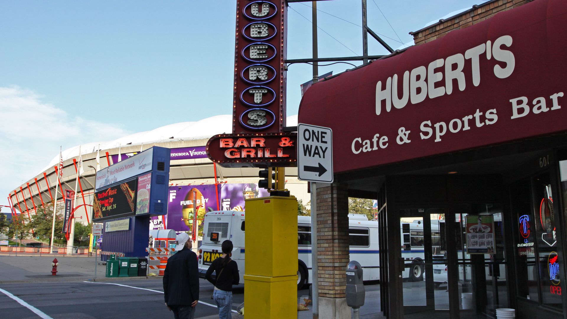 Street scene featuring a tall vertical neon sign reading Hubert's, a red awning that says Hubert's Cafe & Sports Bar, and a glowing Bar & Grill sign; two people walk by under a blue sky.