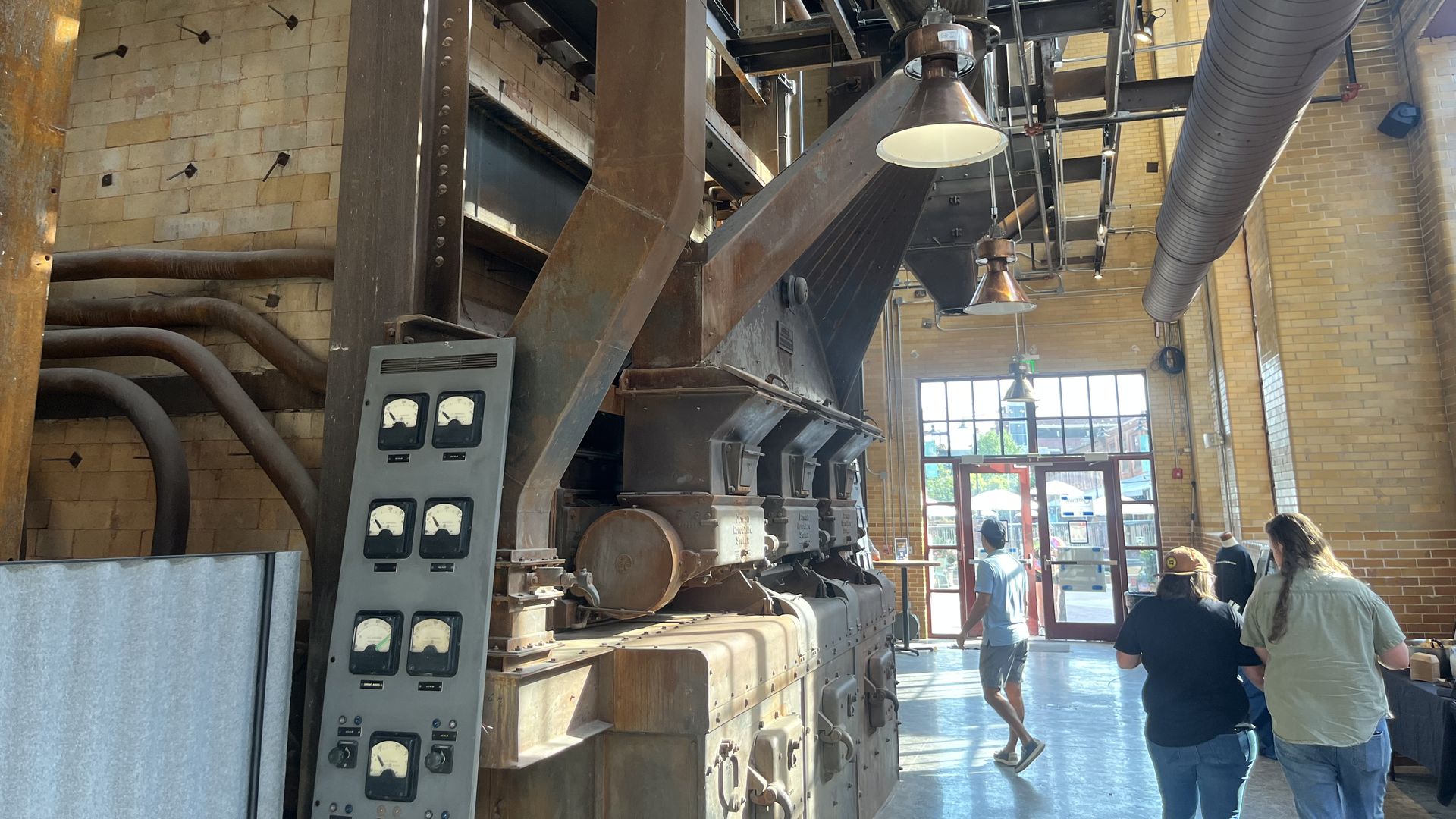 Industrial interior with rusted steel beams, pipes, and a control panel with gauges. A large overhead duct runs along the right; hanging lights, brick walls, and several people near an exit door.