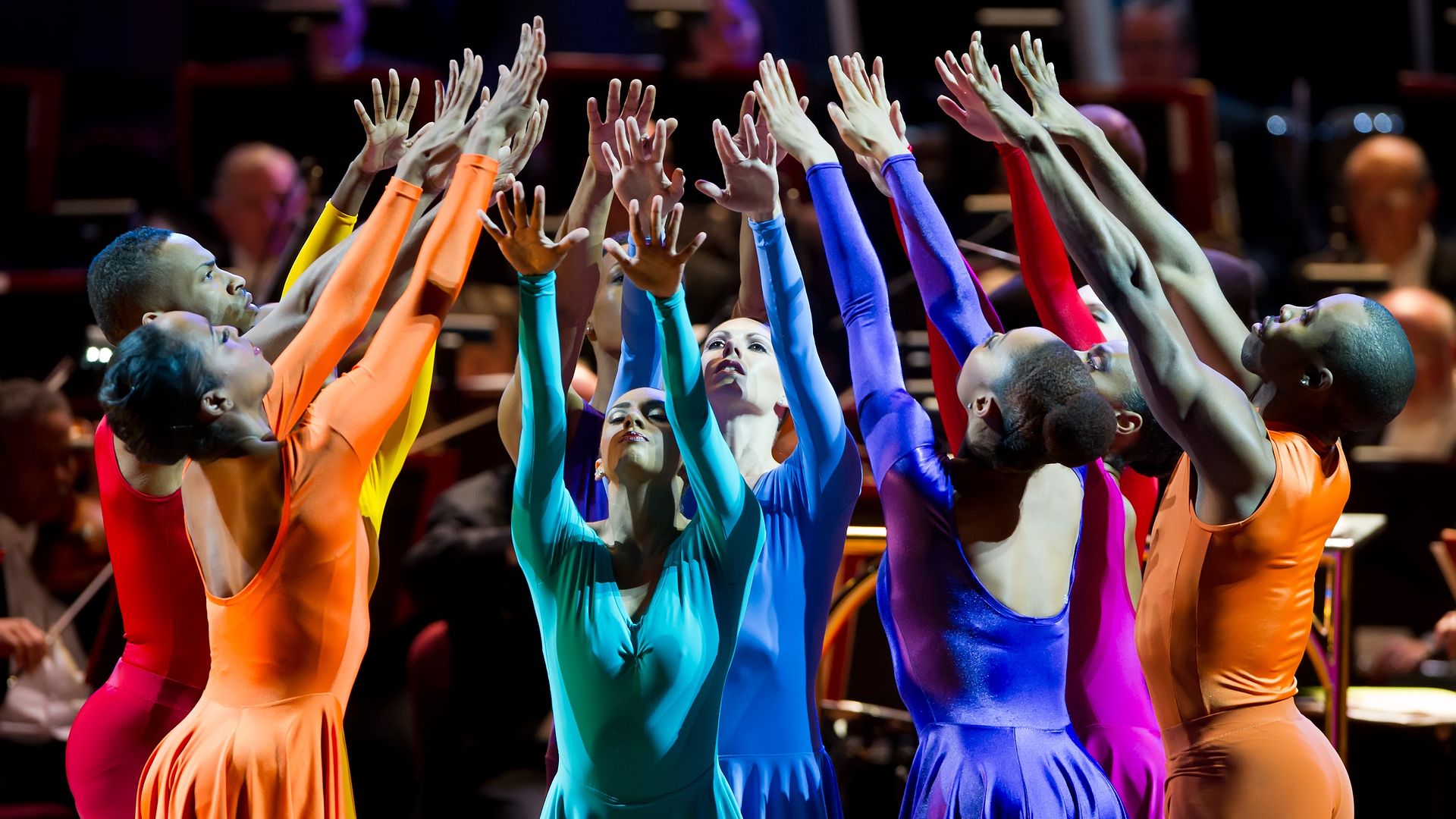 Dancers in brightly-colored costumes stand in a circle with their arms raised.