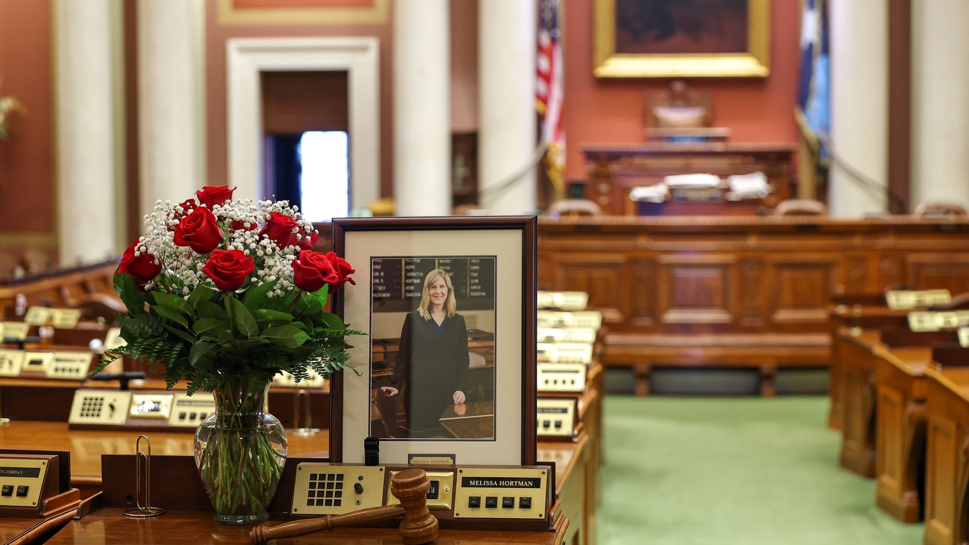 Bouquet of red roses and white baby's breath next to a framed portrait of Melissa Hortman on a desk with a gavel in a wood-paneled legislative chamber with flags and green carpet.