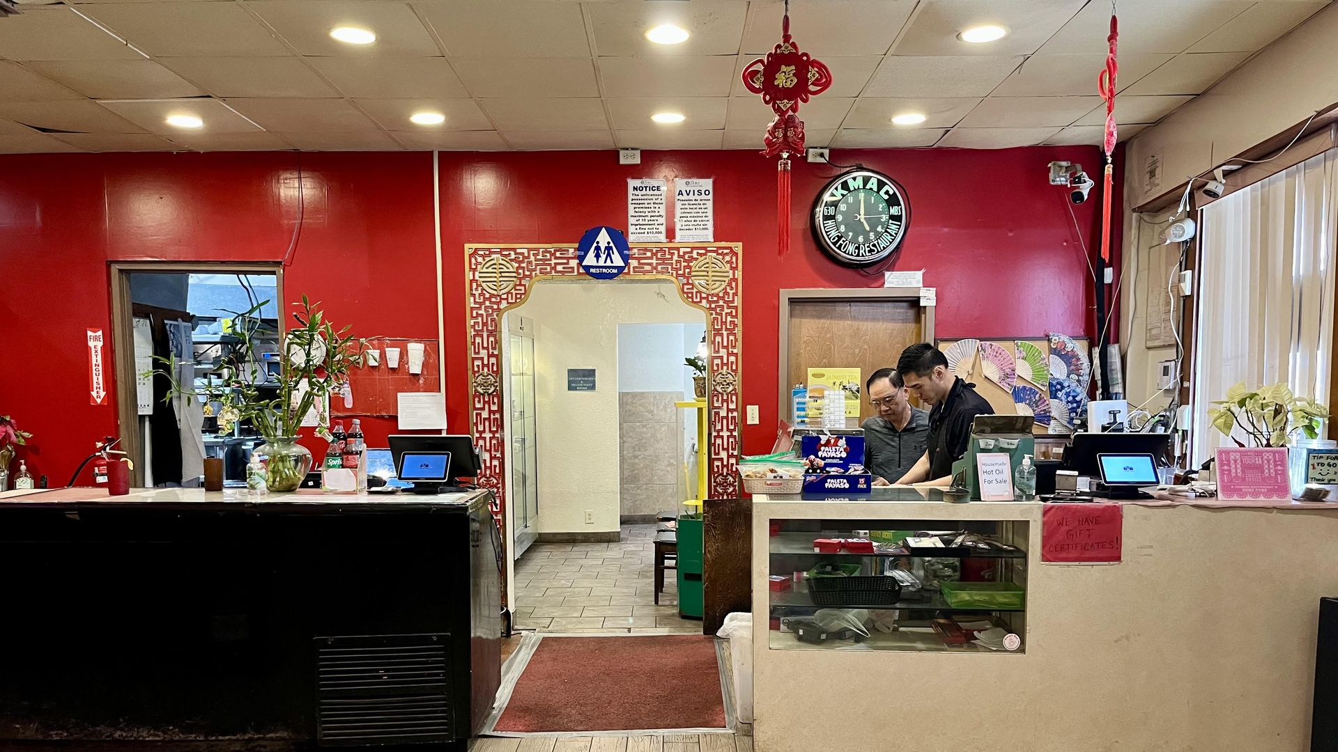 A father and son work at the cash register in front of a bright red wall.