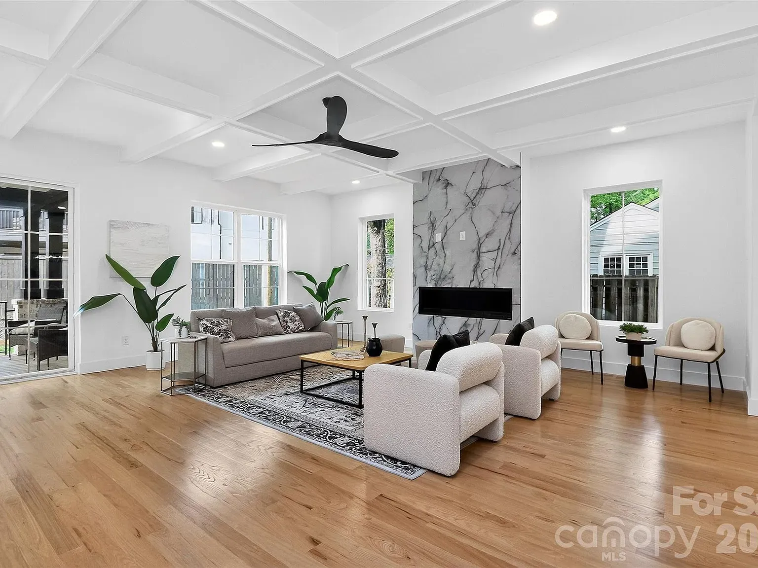 Bright modern living room with light hardwood floors, white coffered ceiling, and a ceiling fan. Gray sofa, white boucle chairs, marble fireplace wall, coffee table, plants, and large windows.
