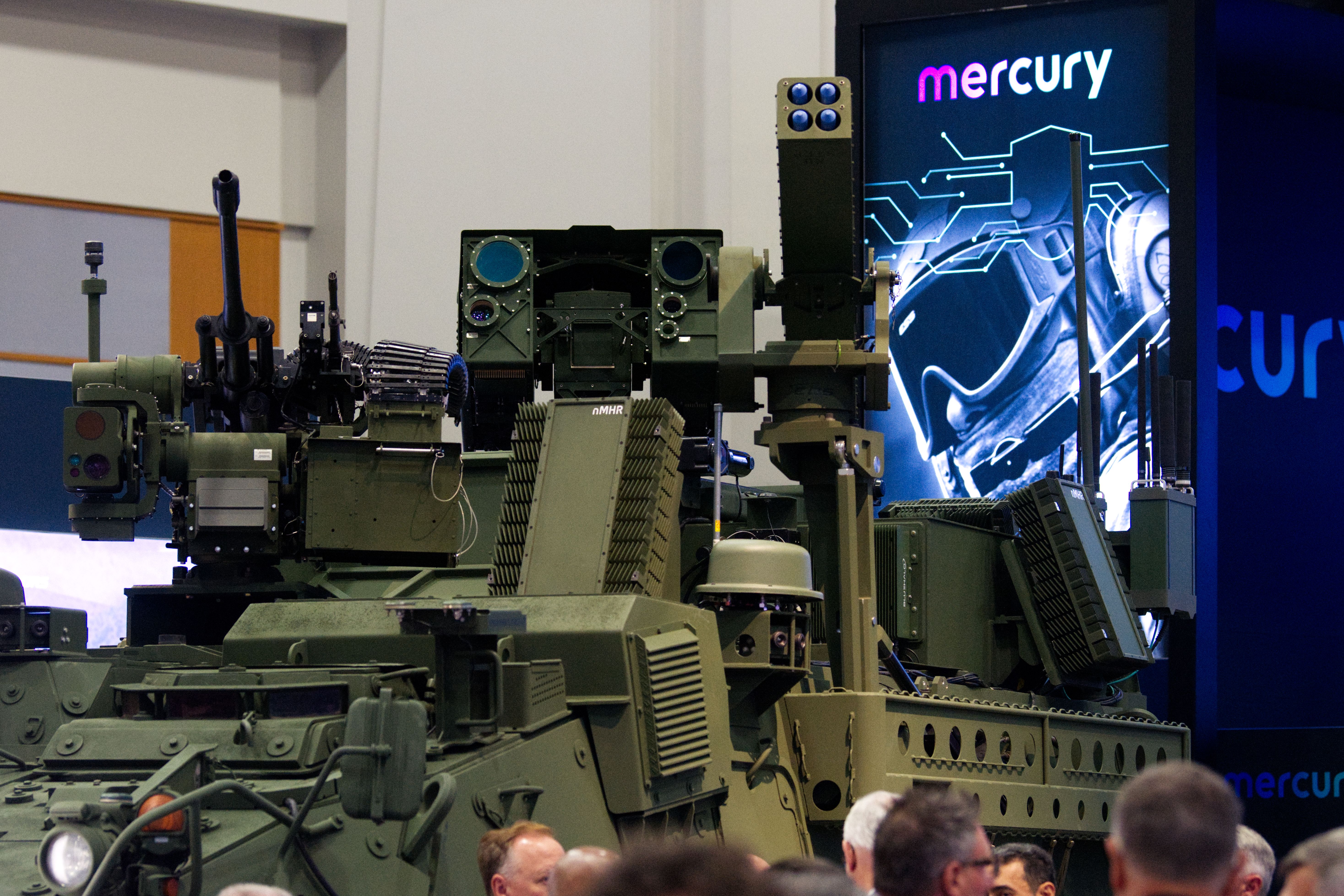 Weapons mounted to a Stryker combat vehicle. People mill about by its wheels. A neon blue sign is seen to the right.