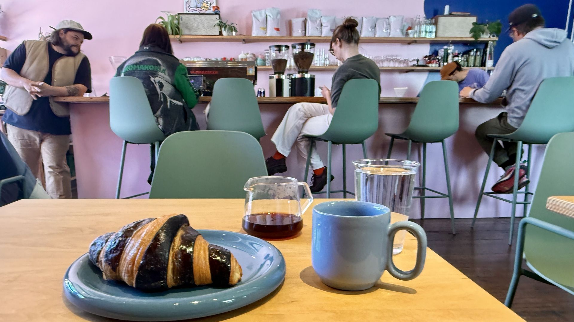 Cozy cafe with pink walls and mint-green chairs along a wooden counter. In the foreground a blue plate with a pastry and a blue mug; several customers sit at the bar, coffee gear on shelves behind.