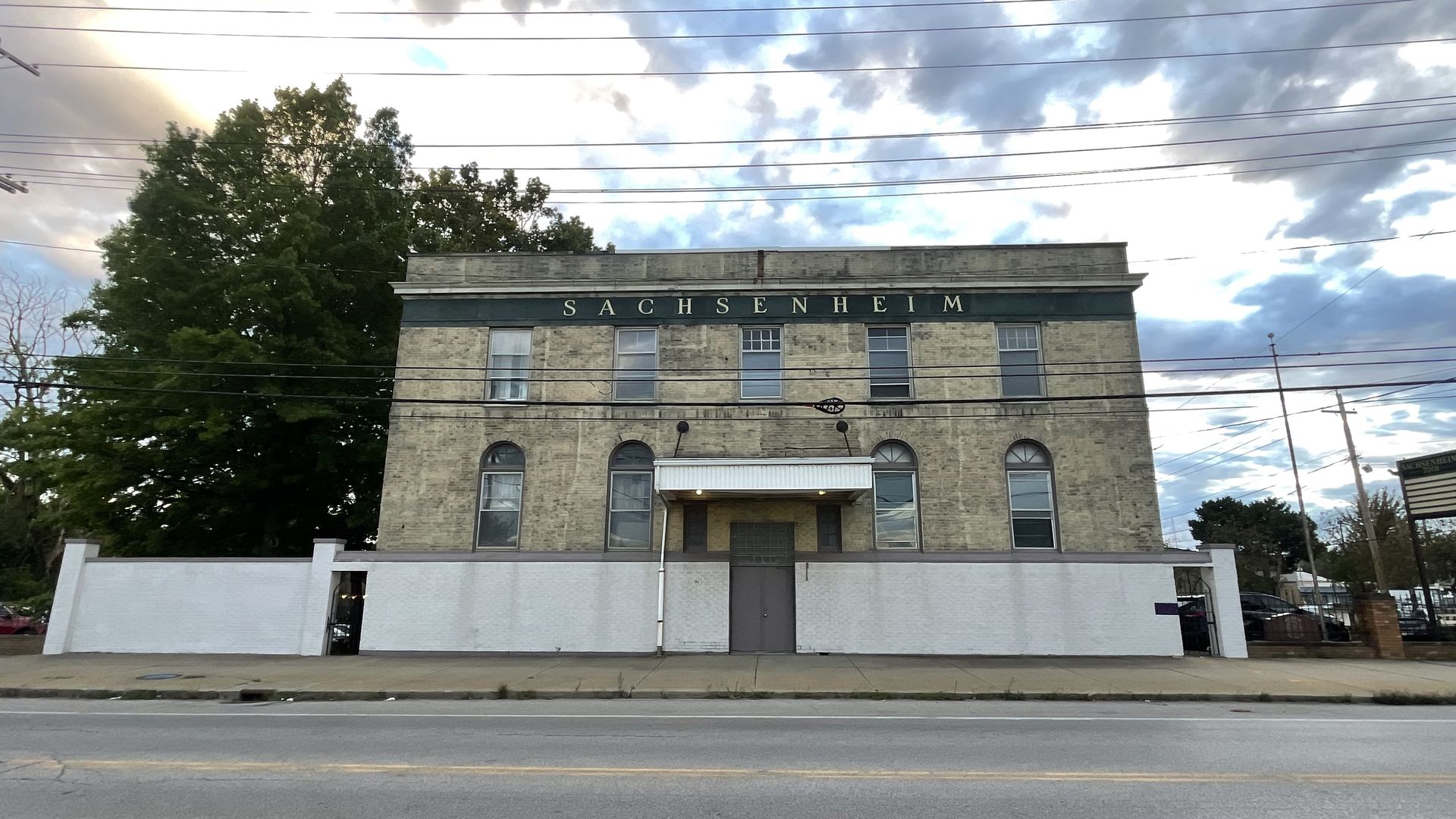 Two-story beige brick building with a blue band at the top spelling SACHSENHEIM. Arched windows, white entry canopy, a large tree to the left, and power lines across a cloudy sky.