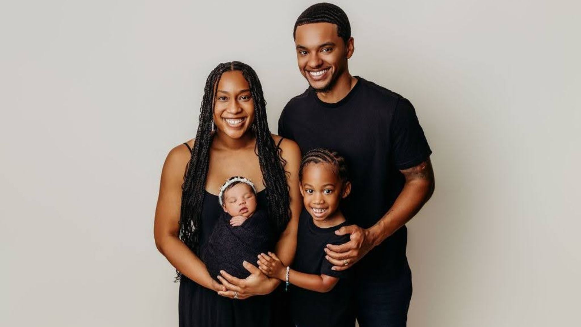 A happy family of four poses against a light gray backdrop: mother with braided hair holds a newborn swaddled in a dark wrap, father smiles beside them, and a young daughter stands in front.
