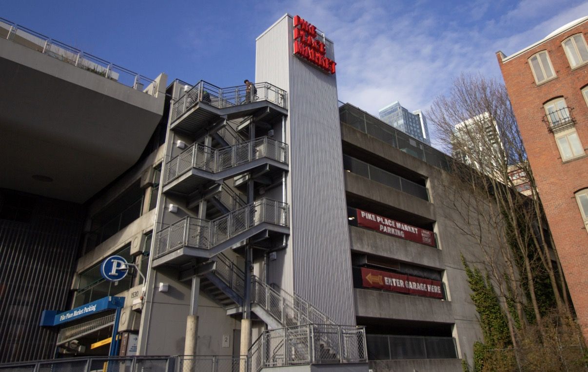 A neon red sign that says "Pike Place Market" is mounted to the side of an elevator tower and parking garage, with a banner that says "Pike Place Market parking" on one side.