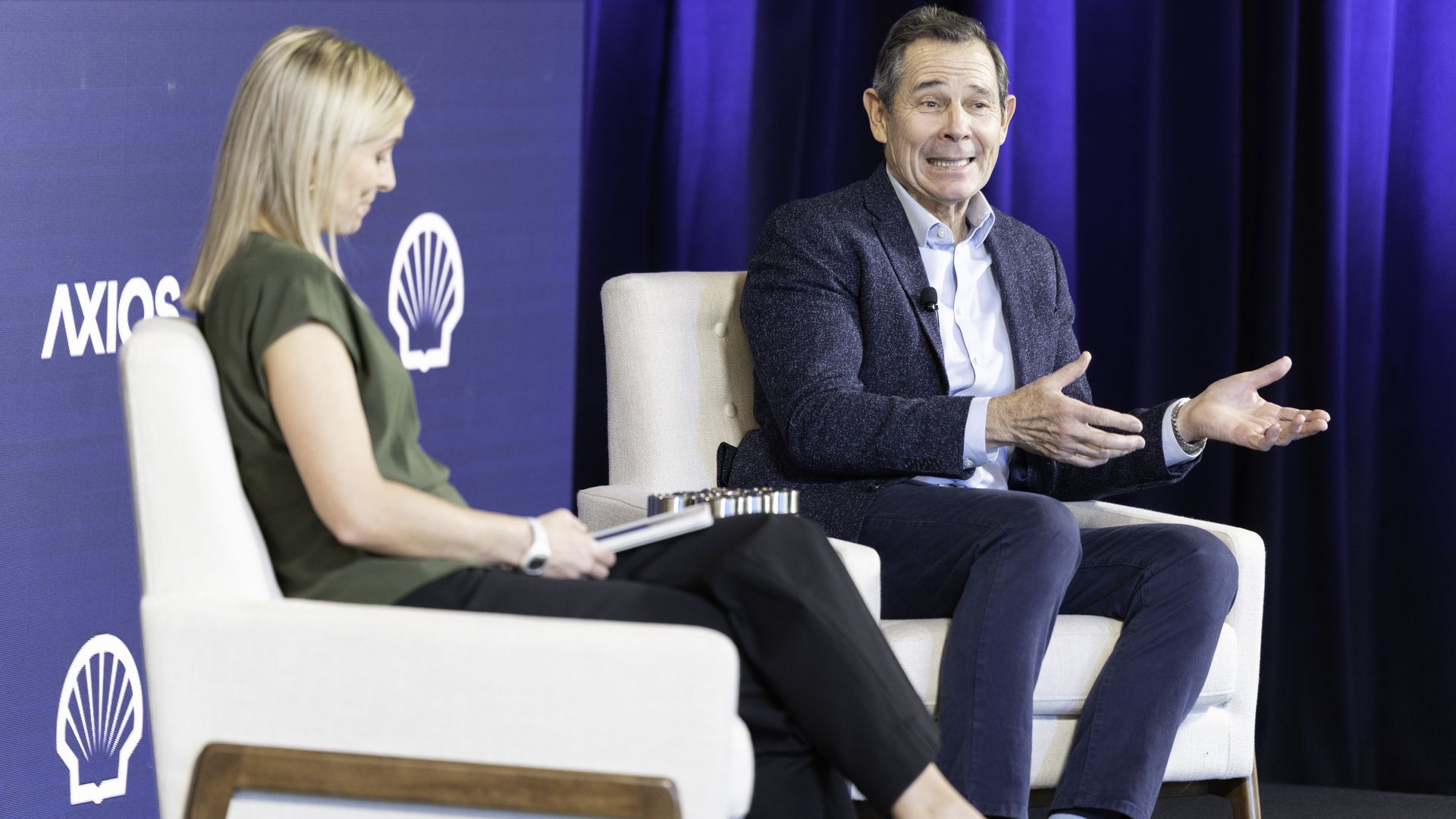 Two people sit in white armchairs on a stage with blue curtains and logos of Shell and Axios behind them. Man on right in blue jacket gestures while speaking; woman on left takes notes.