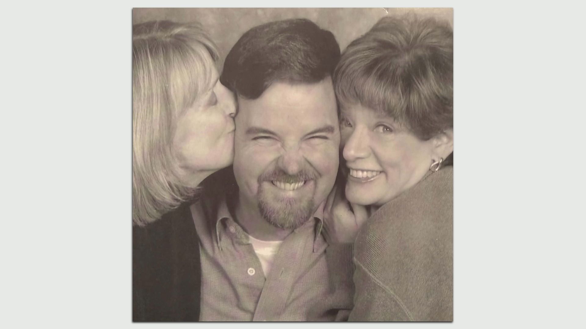 Sepia-toned photo of a smiling man flanked by two women kissing his cheeks; left is a blonde woman, right has short light hair.