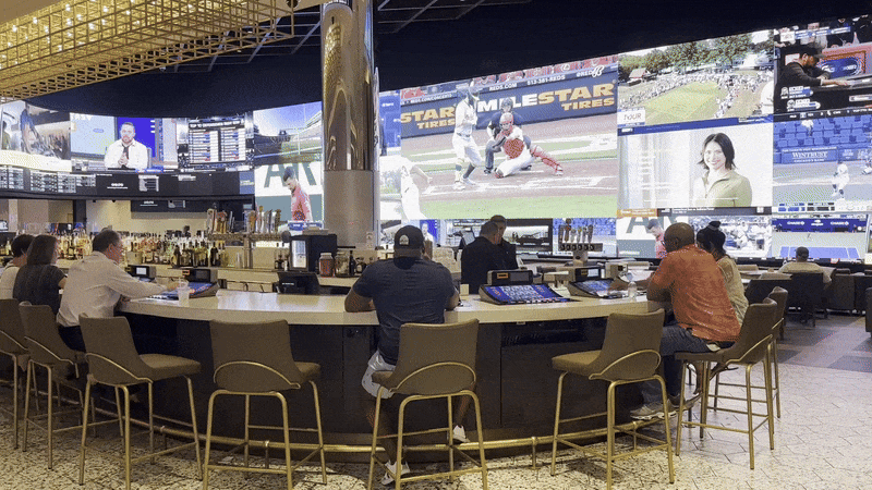 Giant TV screens are on the wall behind a busy bar, where people gamble.