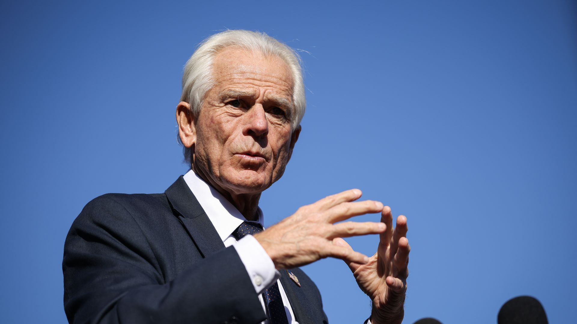 Older man with white hair, wearing a dark suit and tie, speaking and gesturing with hands against clear blue sky, with microphones in the foreground.