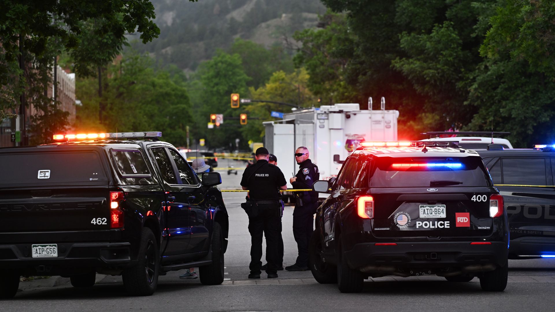 Several police SUVs with red and blue lights line a cordoned city street; officers in dark uniforms stand and talk behind yellow tape, with green trees and distant hills in the background.