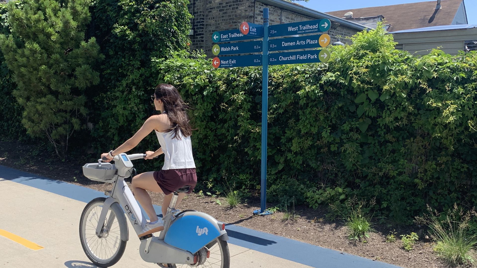 A woman rides a white and blue Lyft bike on a sunny path, beside dense green hedges and a blue signpost with arrows to East/West Trailhead, Walsh Park, and Damen Arts Plaza.