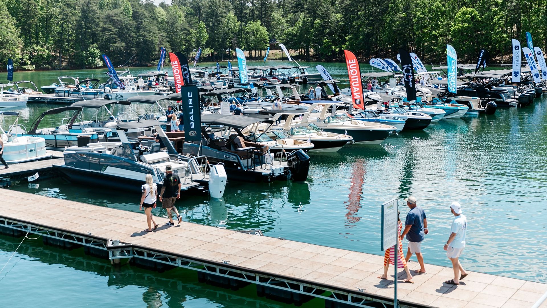 A busy marina with numerous boats docked along a wooden pier, colorful flags fluttering, calm greenish-blue water, and a tree-lined shore under a sunny sky.