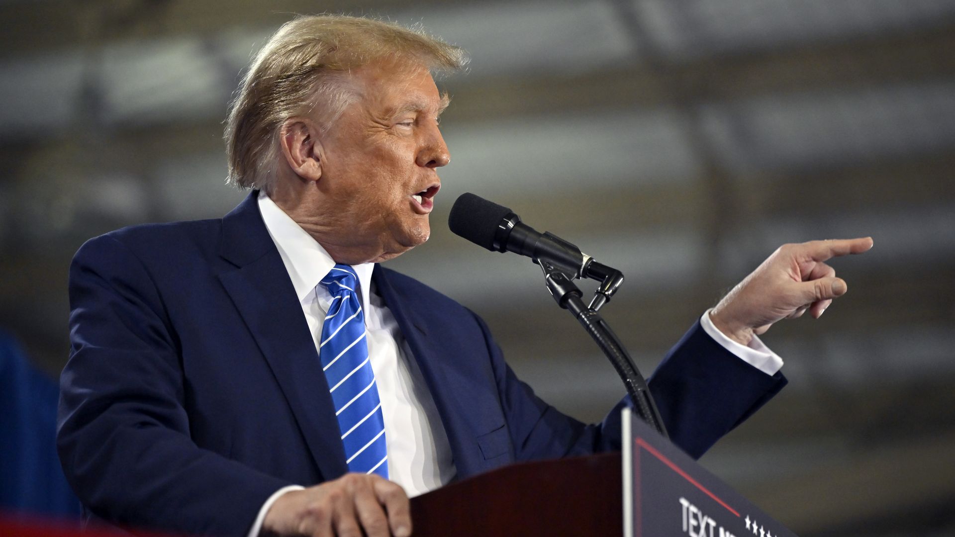 Donald Trump, wearing a blue suit, white shirt and blue tie. points at someone from behind a podium.