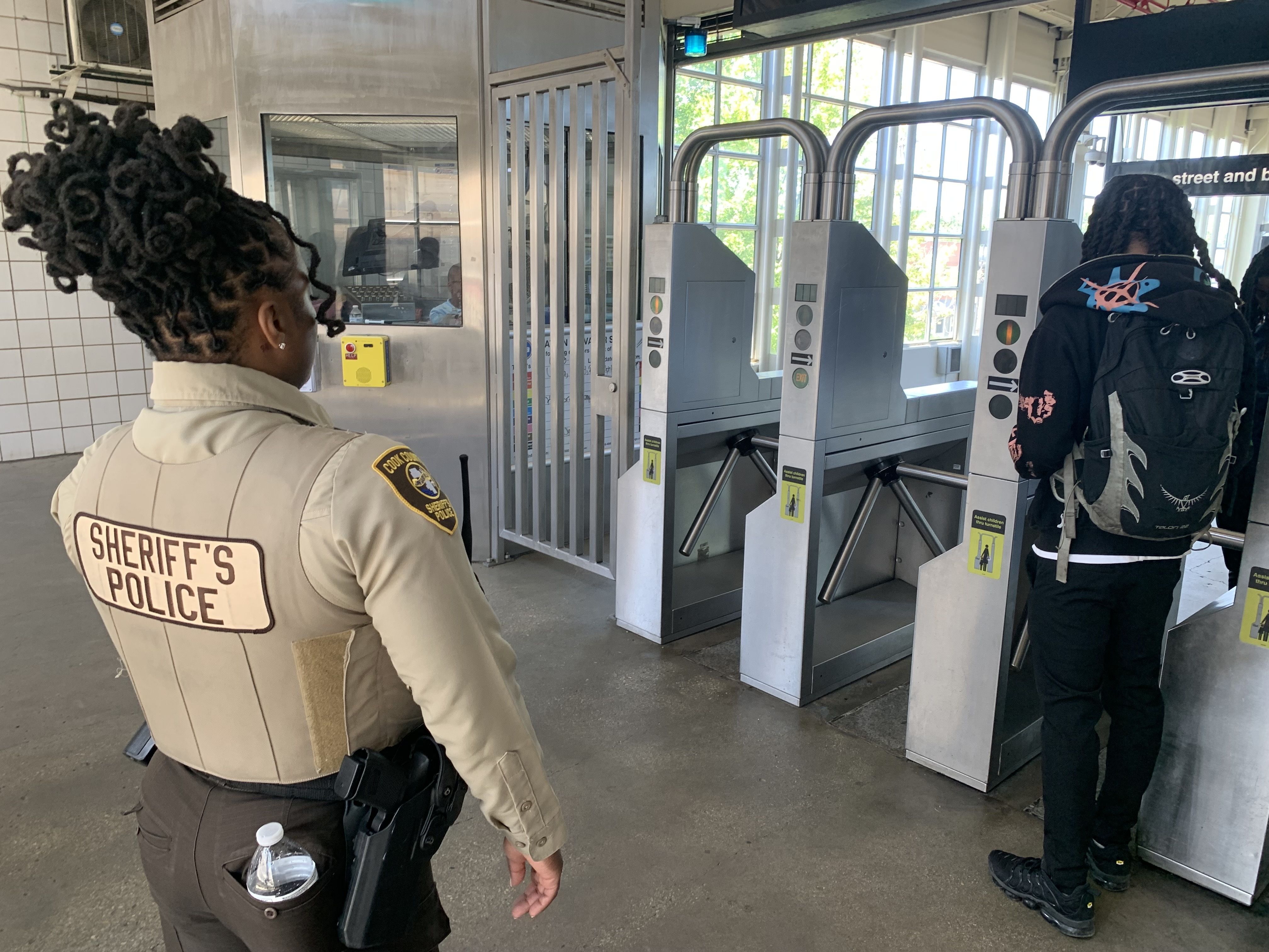 Sheriff's deputy in a beige uniform stands near a row of gray security turnstiles at a station entrance; another person with a backpack waits by large windows.