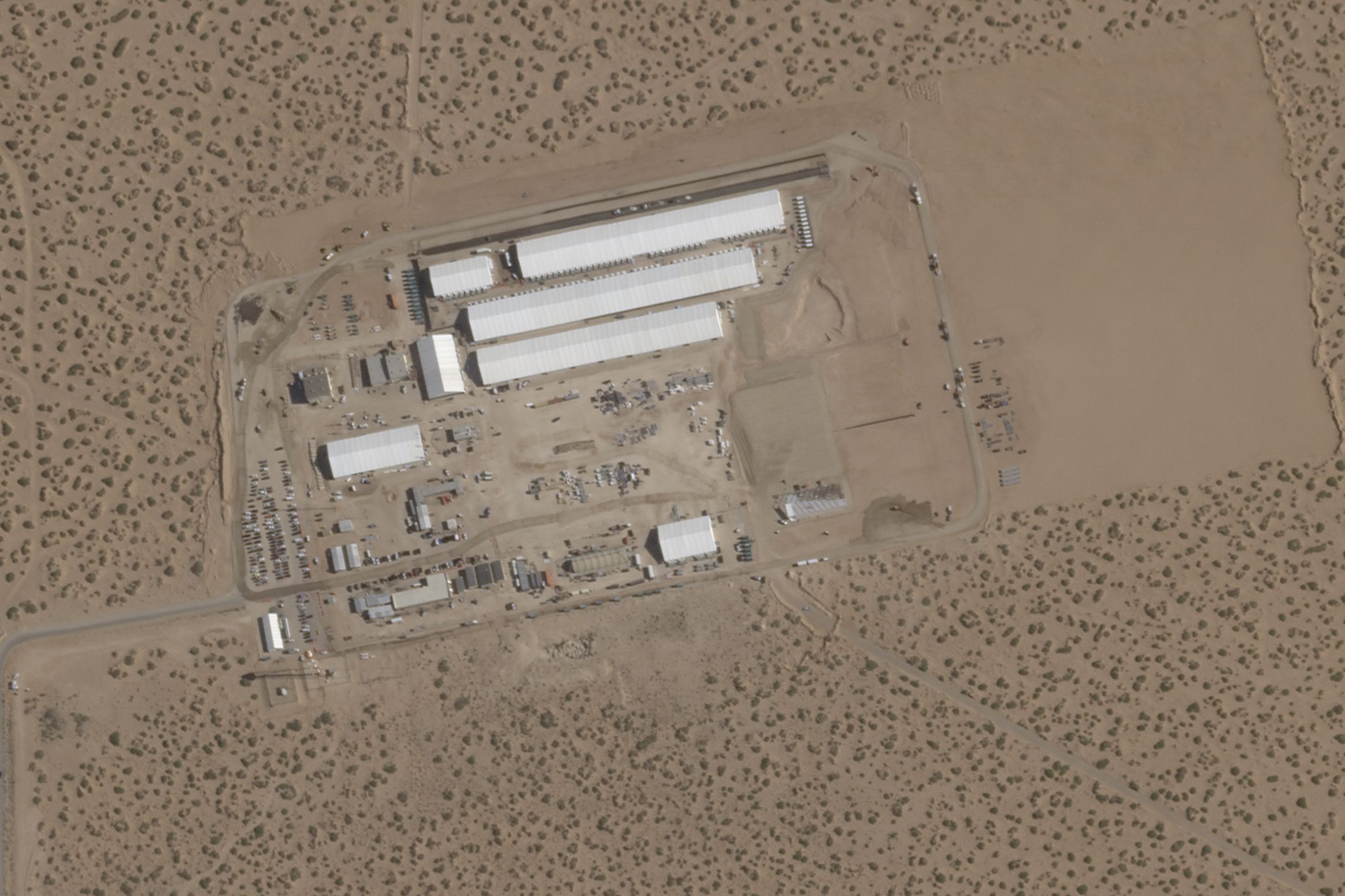 Aerial view of a desert area with multiple long white-roofed buildings arranged in rows, surrounded by dirt roads and sparse vegetation.