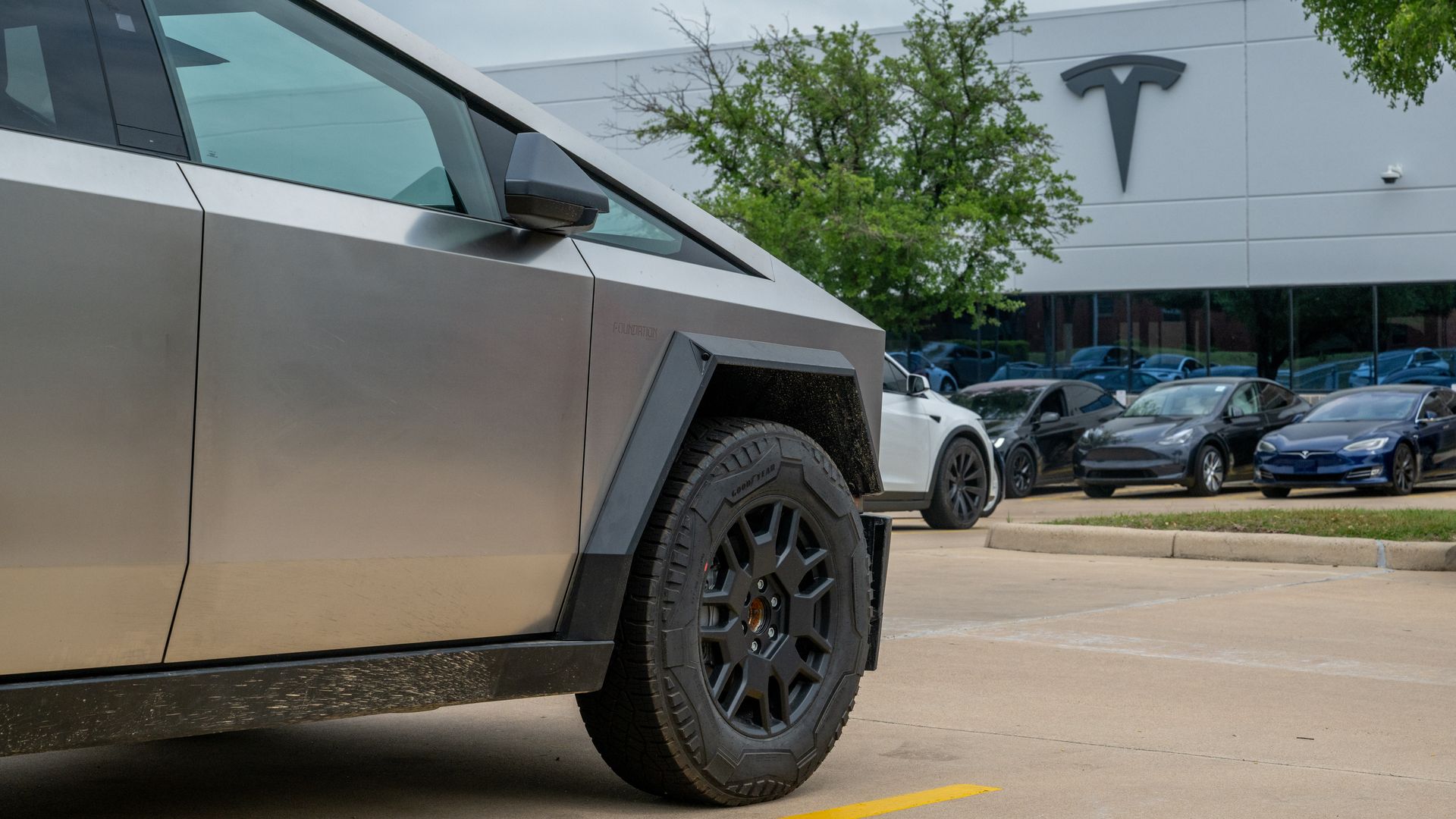A Tesla Cybertruck sits in a parking spot of a Tesla dealership with a Tesla logo in the background.