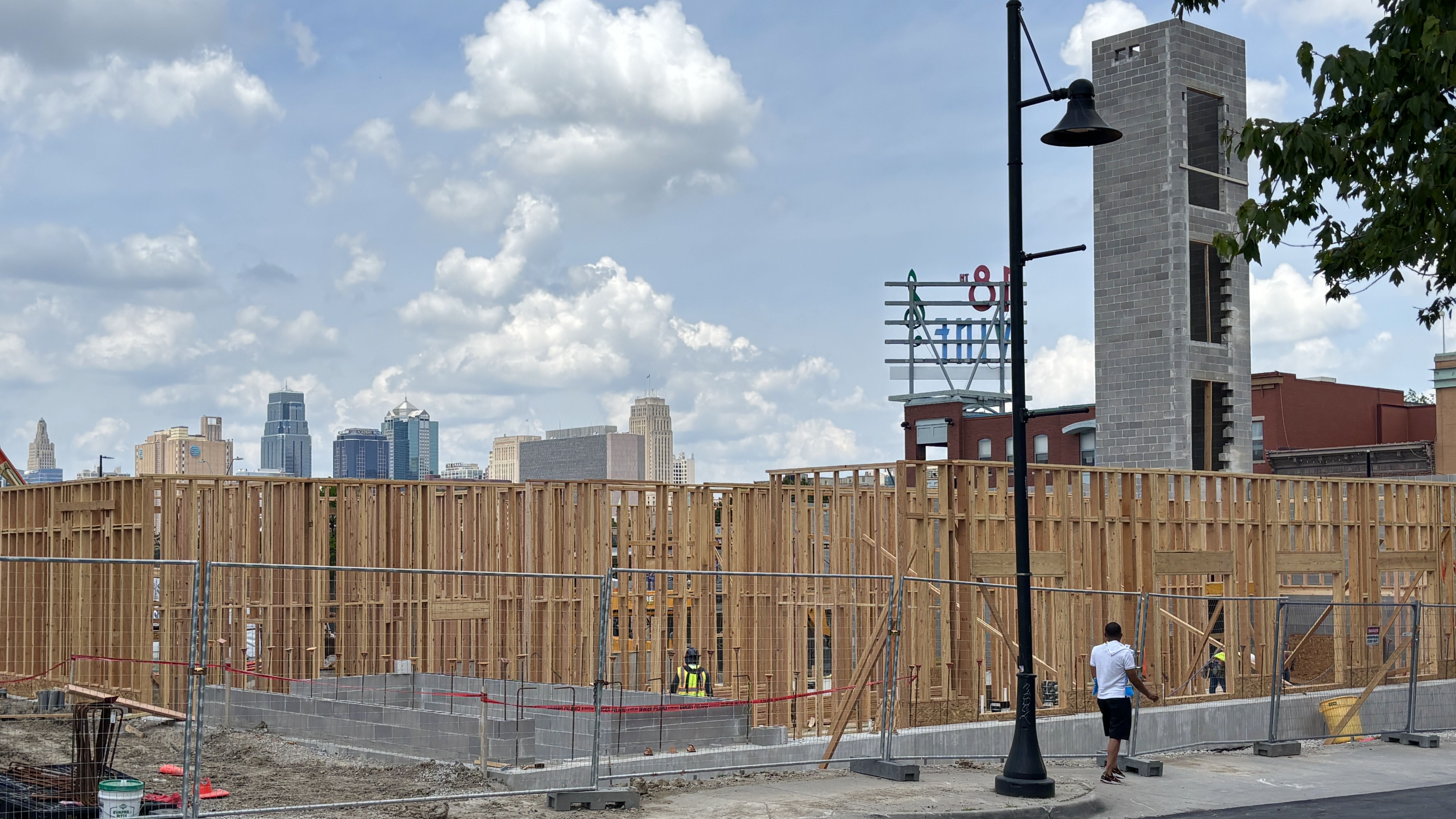 A construction site with wooden framing in the foreground, a tall gray cinder-block tower, a black streetlamp, and a distant city skyline beneath a blue sky with white clouds.