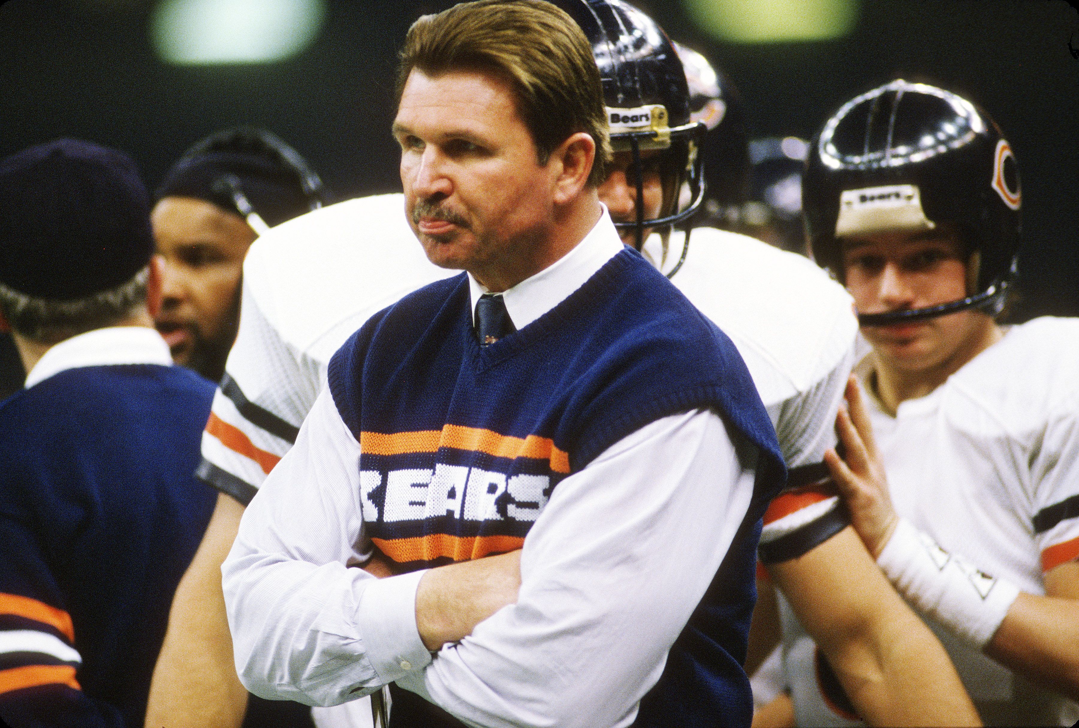 Man with mustache and slicked hair wearing Chicago Bears sweater vest over white shirt with arms crossed, football players in helmets and white uniforms behind him.