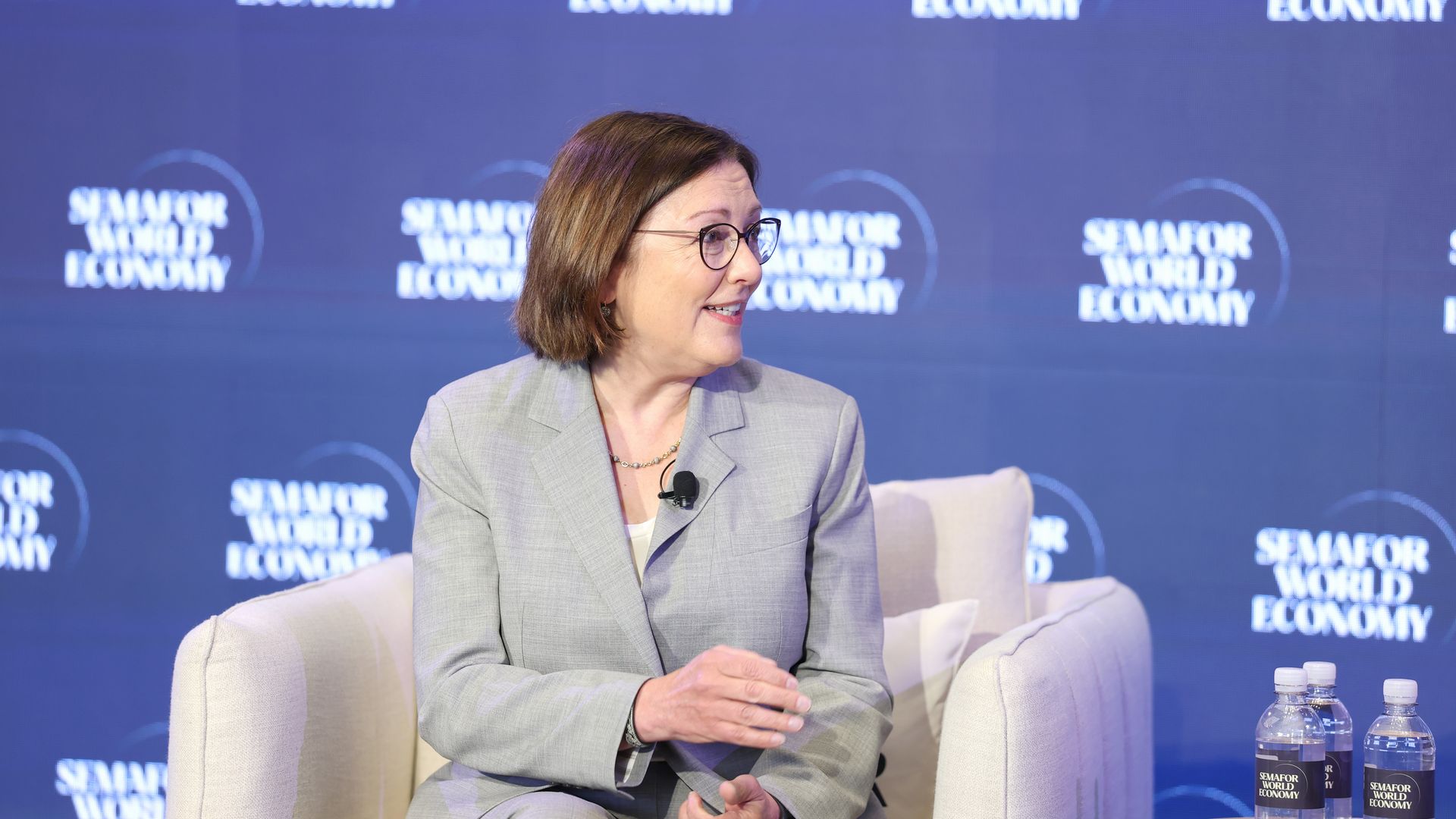A woman with short brown hair and glasses sits on a stage in a gray blazer, against a blue backdrop with circular logos and the text "SEMAFOR WORLD ECONOMY"; a microphone on her lapel and water bottles nearby.