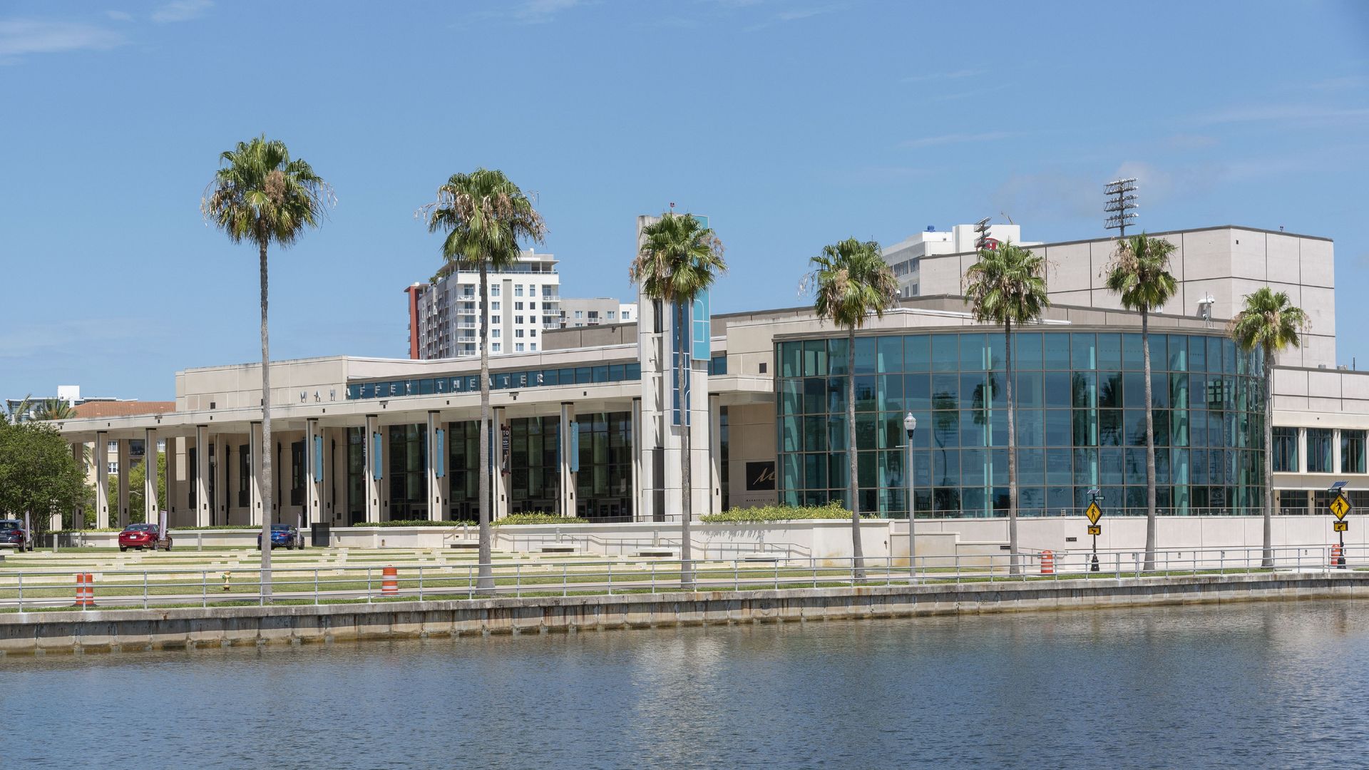 Sunny day at a waterfront campus: beige glass-front building with palm trees along a promenade, calm water in the foreground, blue sky, and a few parked cars.