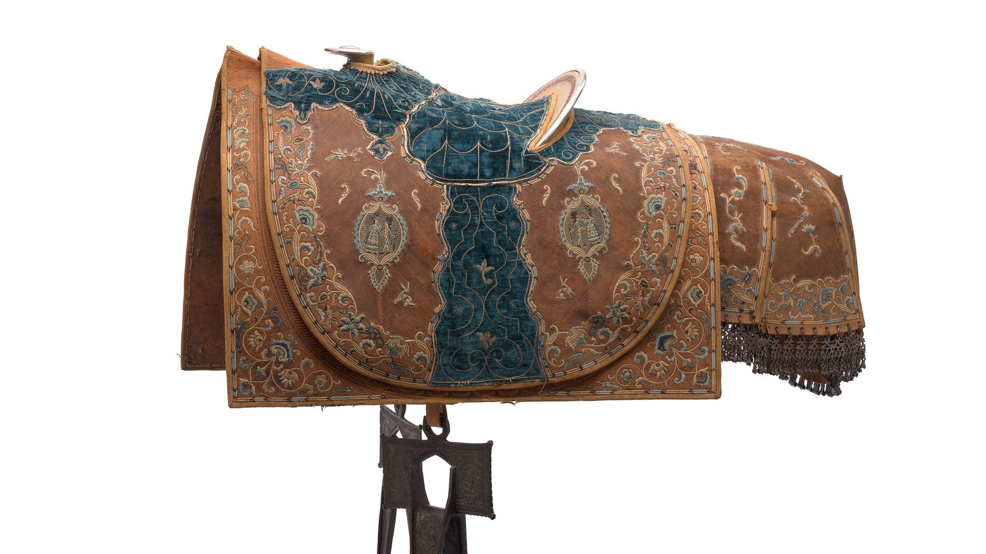 Ornate antique saddle on a stand, brown and teal embroidered fabric with floral patterns, silver scrolls, hanging stirrups, and fringe along the edge.