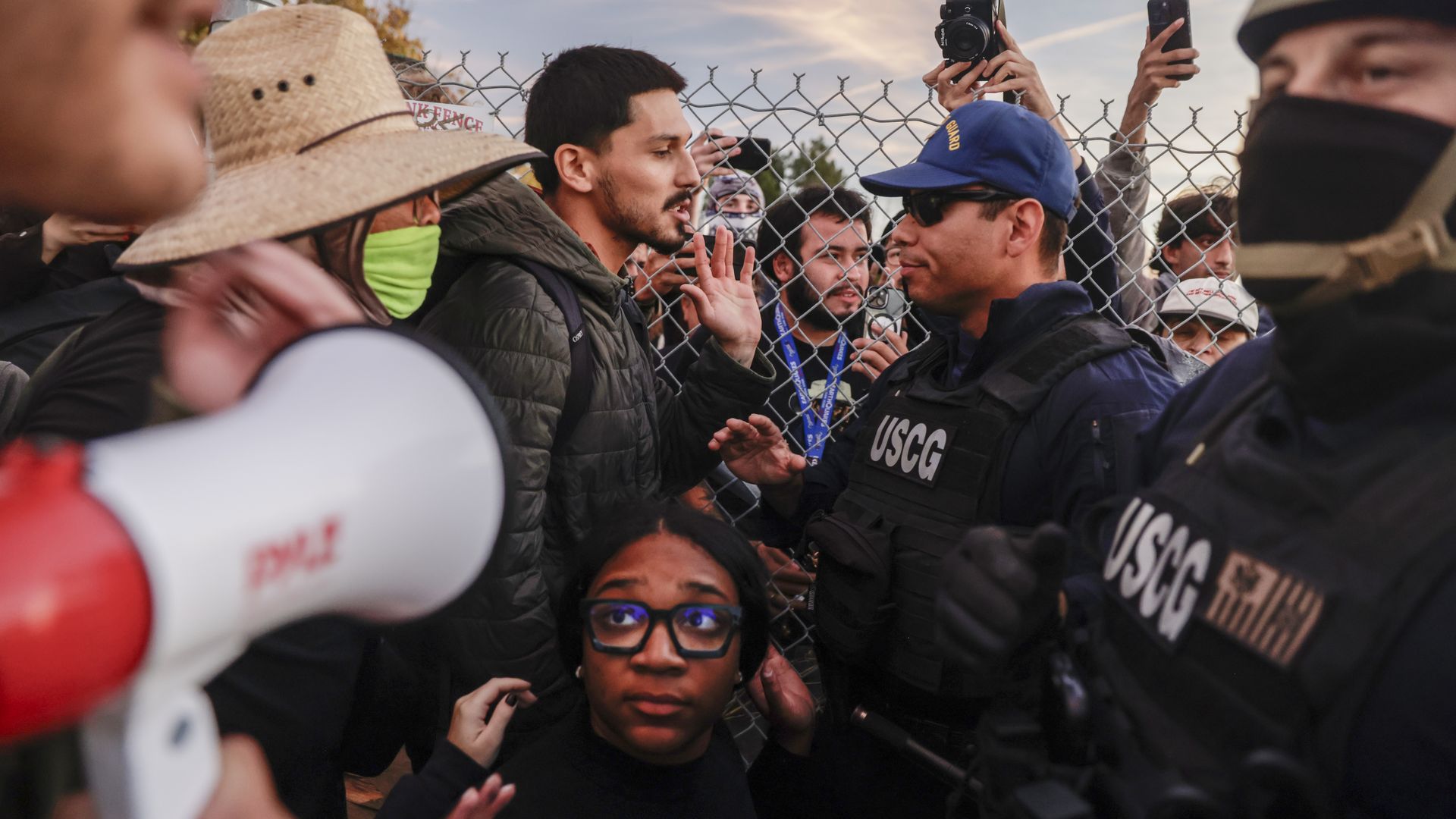 People, including a man and a woman, engage with USCG officers in dark uniforms near a chain-link fence. Someone holds a megaphone, and others take photos and videos with phones.