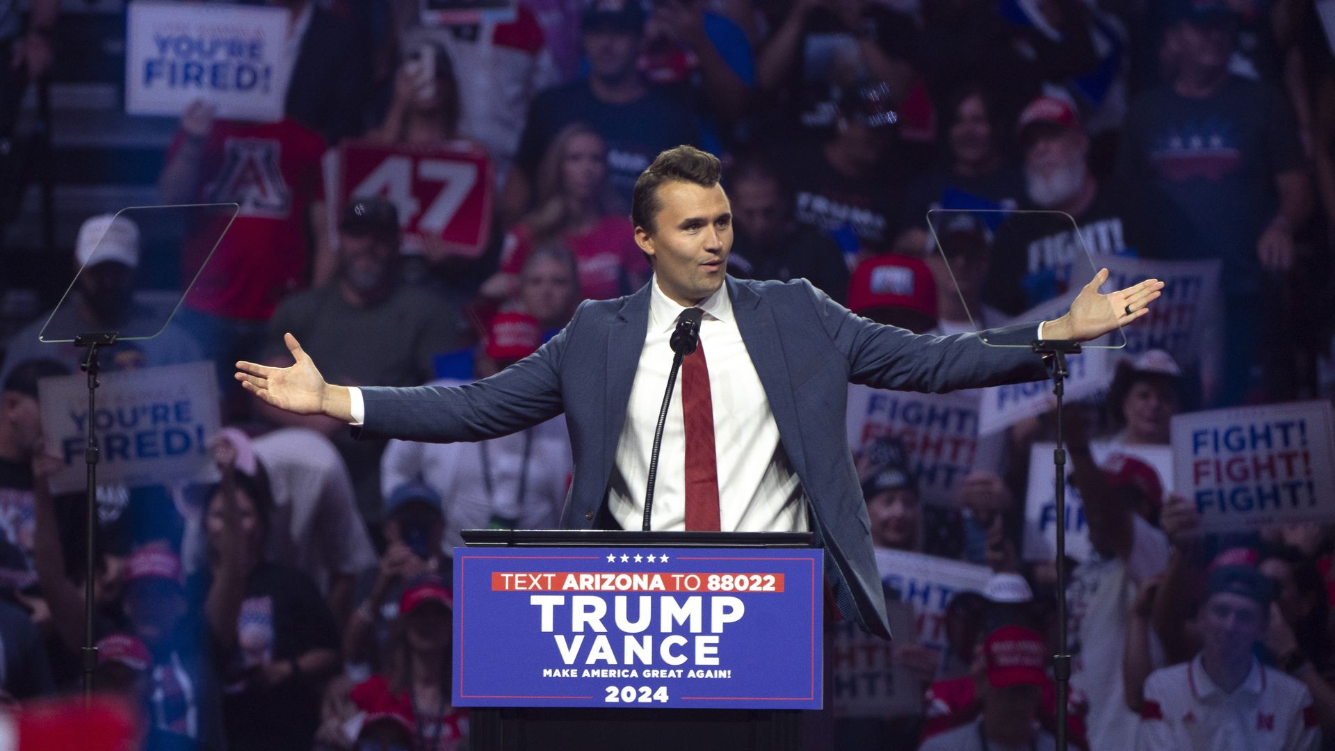 Charlie Kirk, wearing a blue suit and red tie, raises his arms as he speaks into a microphone attached to a podium that reads "TRUMP VANCE."